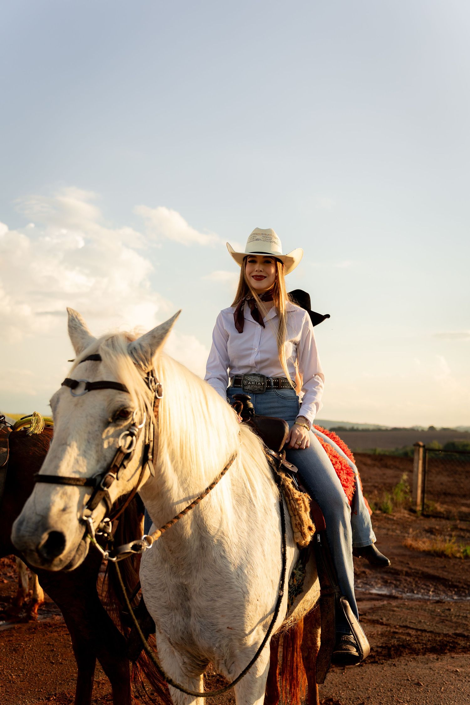 Smiling young woman in white cowboy hat and jeans riding a white horse outdoors in golden hour light.