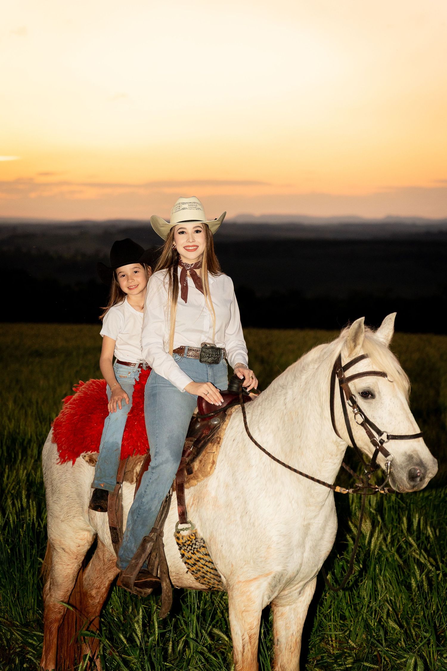 Two women in cowboy hats ride a white horse through a green field at golden sunset.