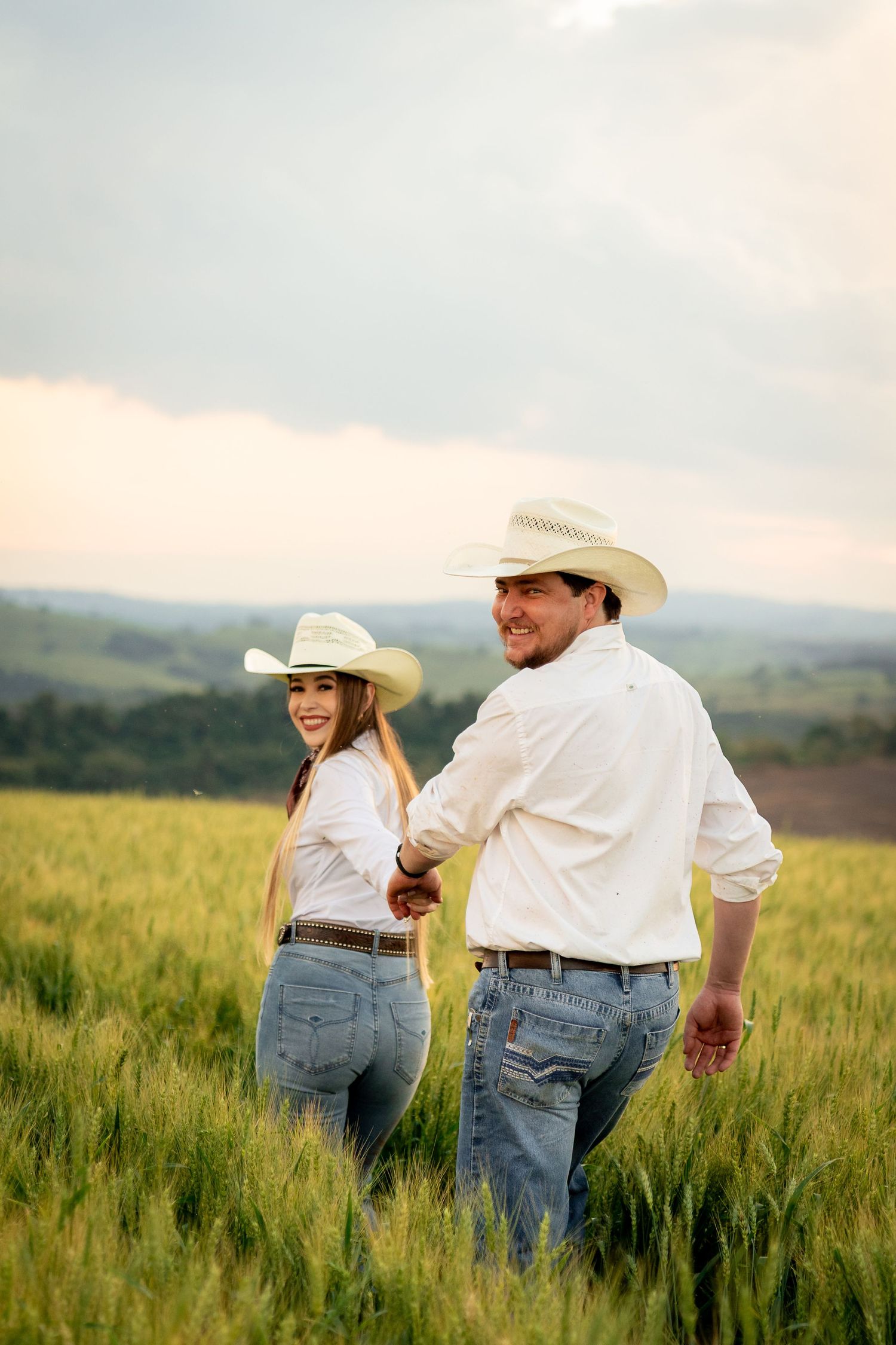 A couple in matching white shirts, jeans, and cowboy hats hold hands while walking through a green field.