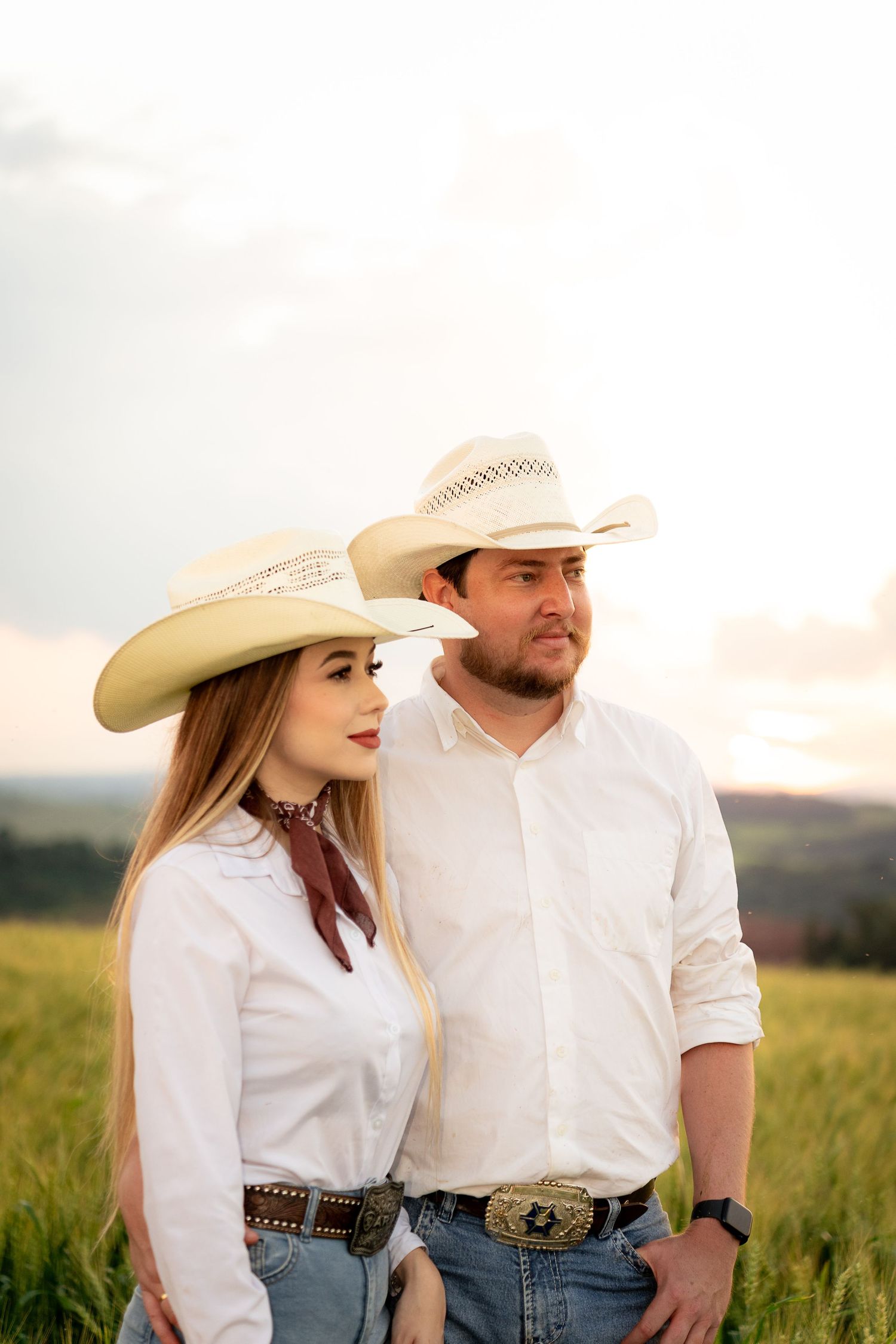 A couple wearing matching white shirts and cowboy hats stand together in a green field at sunset.