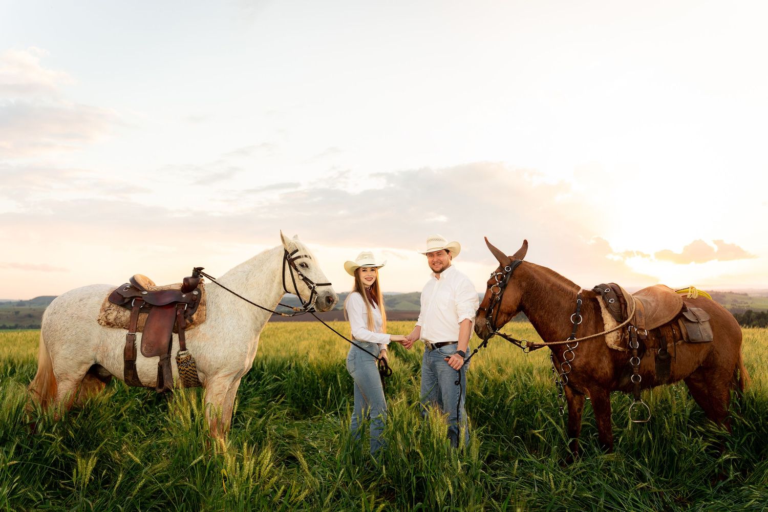 Couple in cowboy hats holding horses in a lush green field at sunset.
