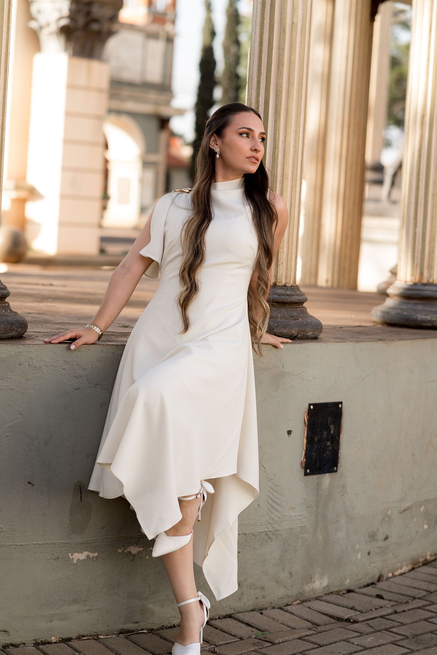 Young woman in elegant white midi dress with asymmetric hem poses gracefully by classical columns in golden light.