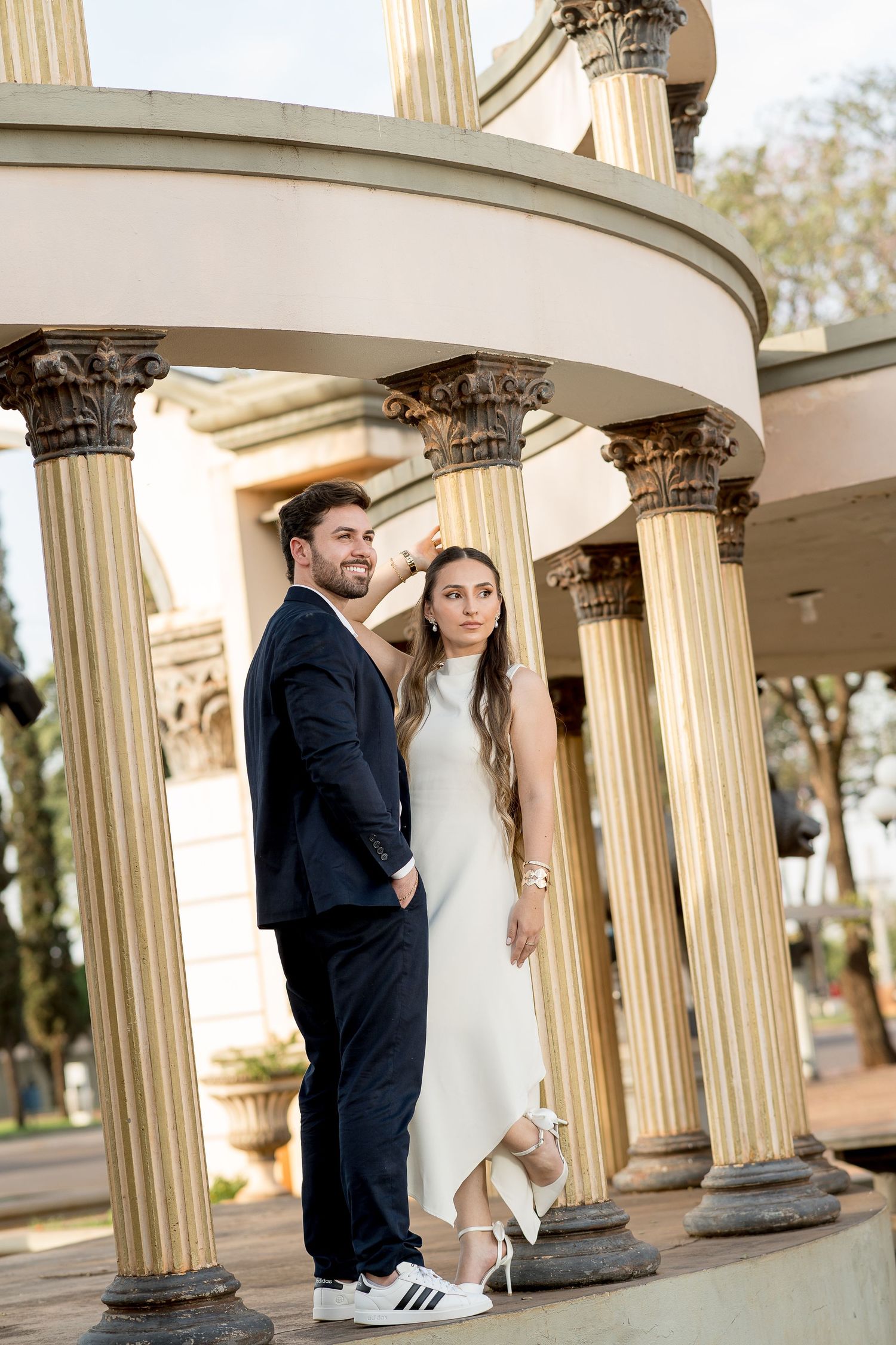 Elegant couple posing under classical columns, man in navy suit and woman in white dress at an outdoor architectural landmark.