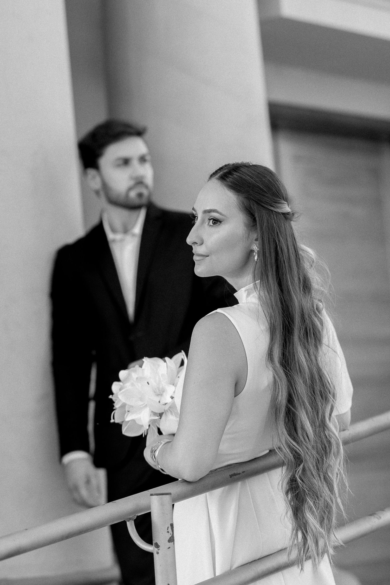 Black and white wedding photo of bride holding bouquet in white dress with groom in dark suit standing behind her.