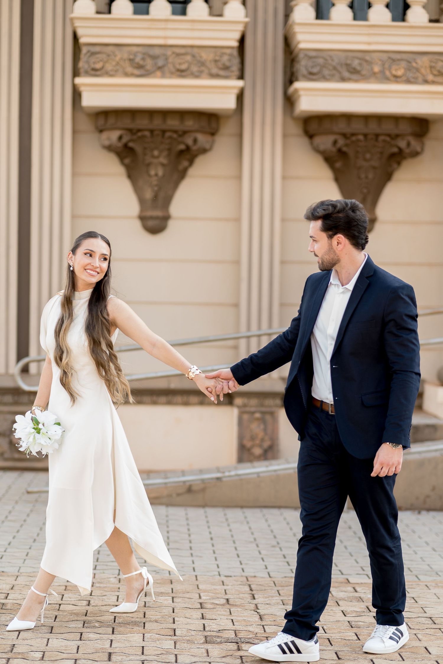 Bride in white dress holds groom's hand in navy suit, walking past ornate building facade on their wedding day.