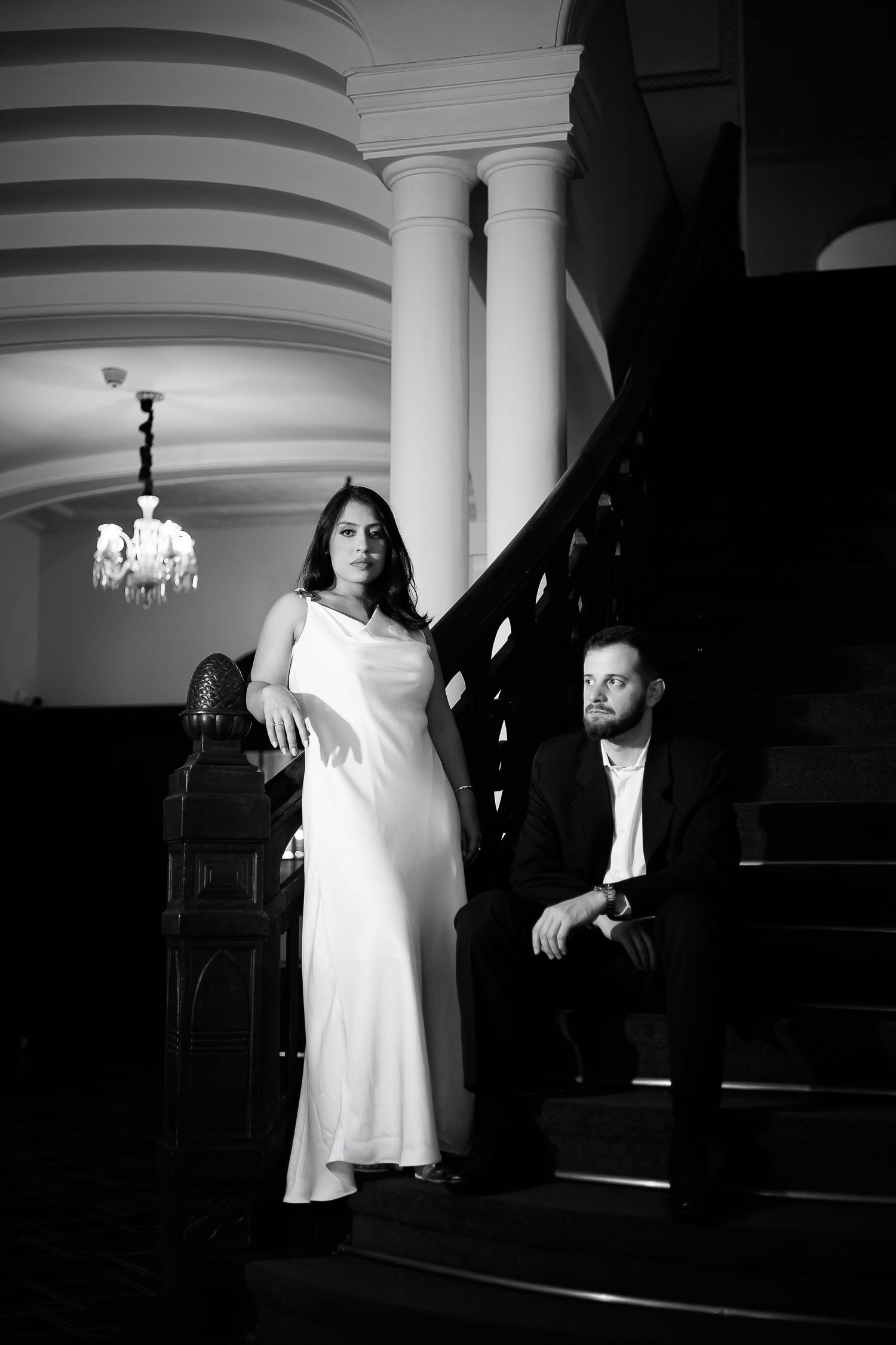 Elegant black and white photo of a woman in white gown and suited man posed on grand staircase with chandelier.