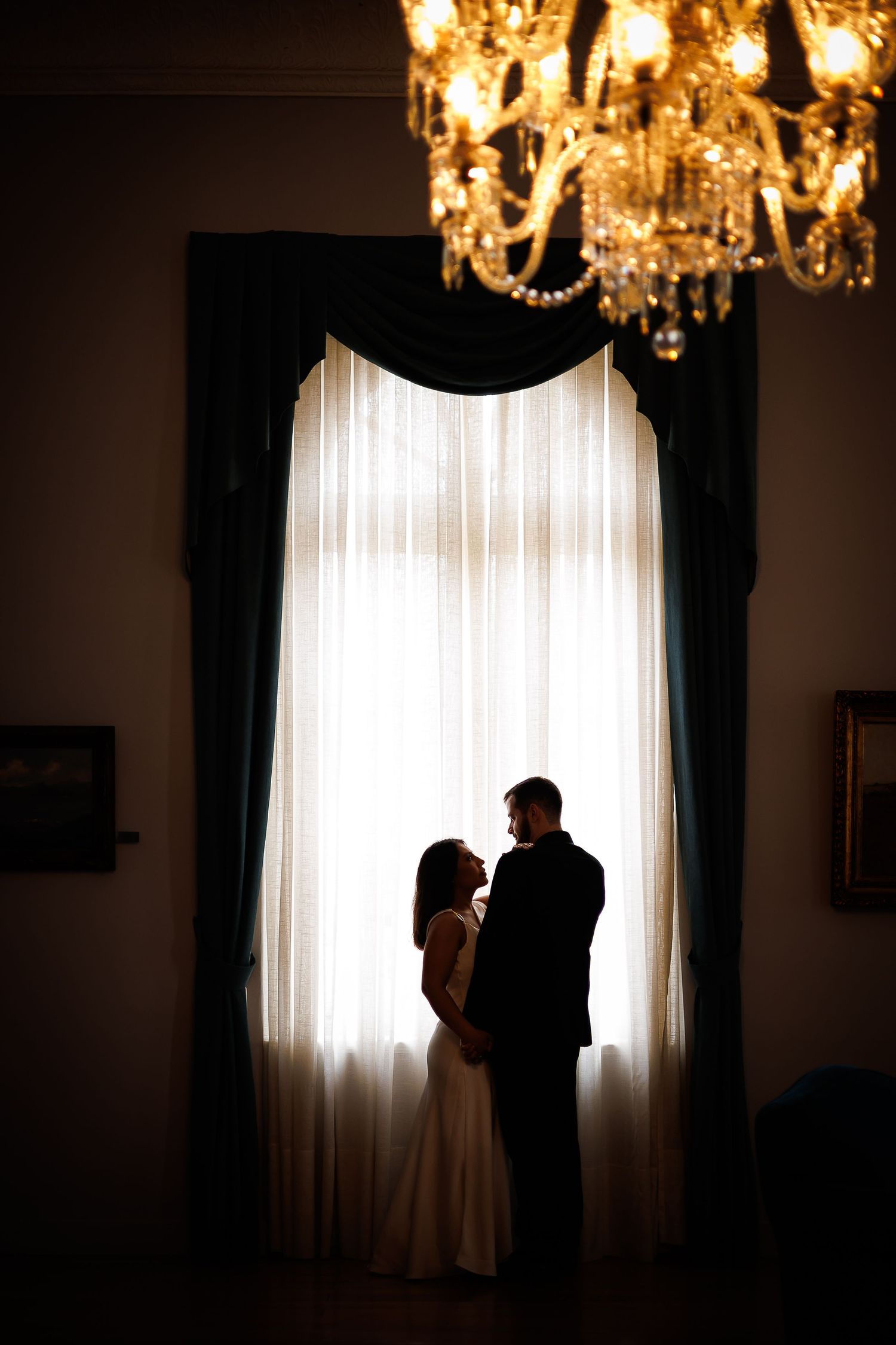 Romantic silhouette of a couple standing before a grand window with teal drapes, lit by an elegant crystal chandelier.