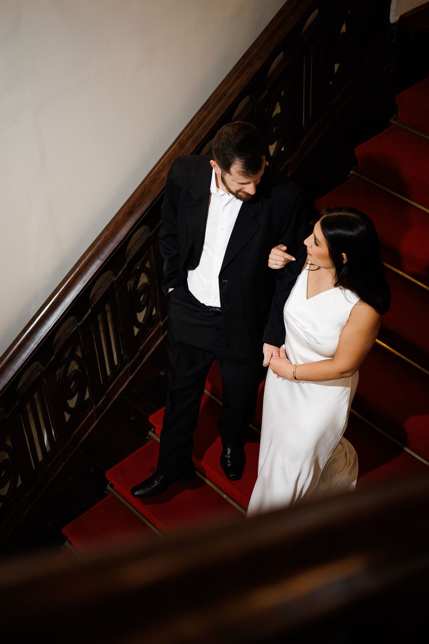 Bride and groom share a romantic moment on a grand staircase with red carpet and dark wood railings.