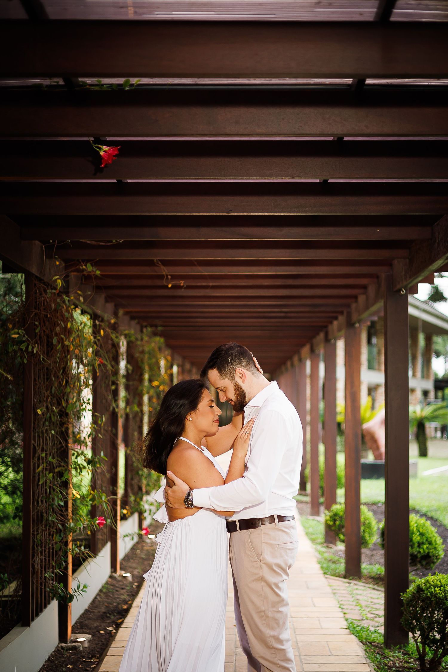 Couple embracing romantically under a wooden pergola surrounded by lush greenery, wearing white wedding attire.