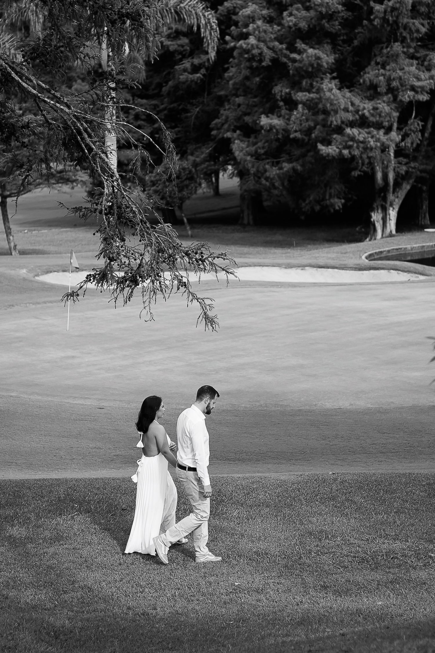 Black and white photo of a couple in white attire walking hand in hand on a lush golf course with trees.