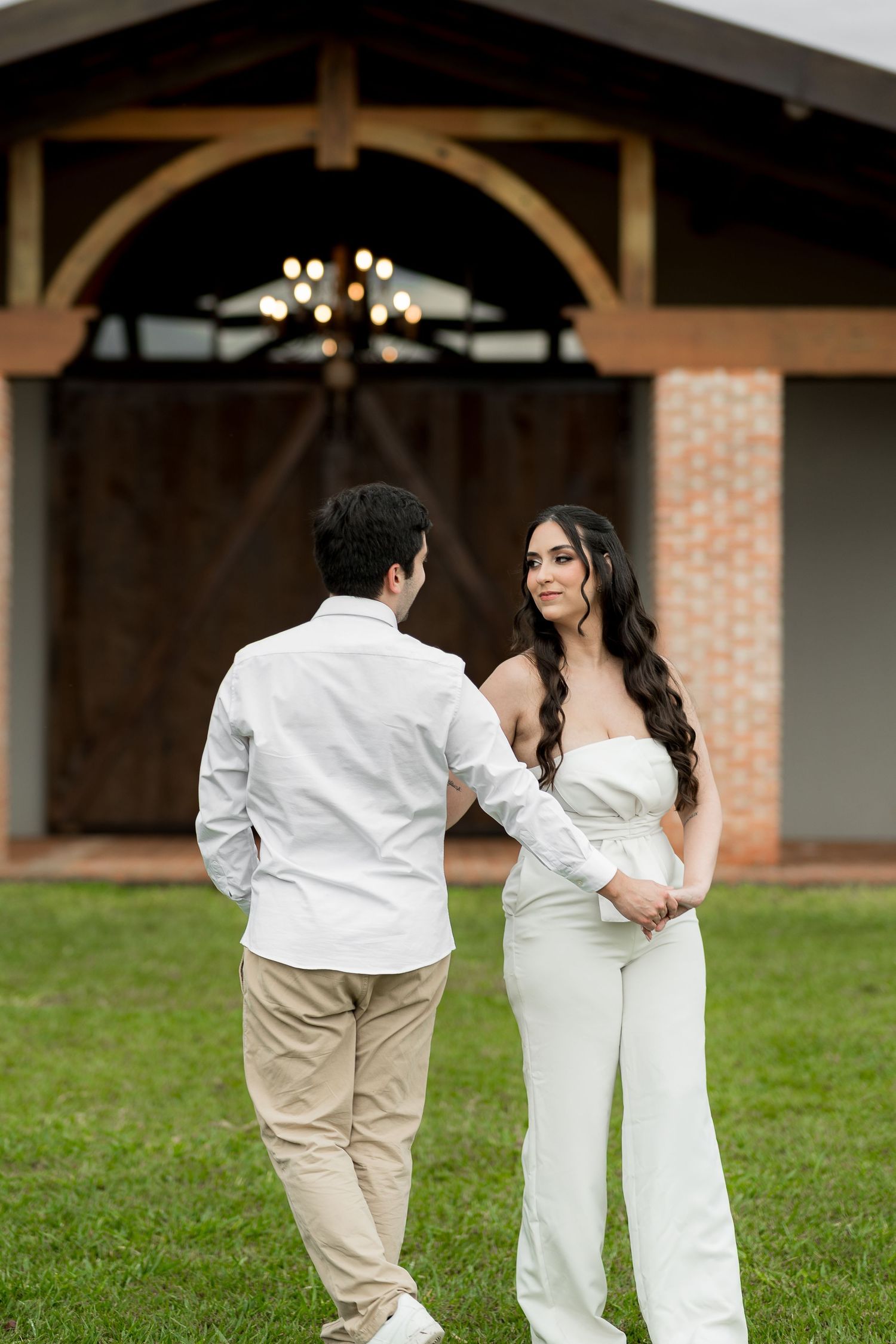 Couple in white outfits holding hands on green lawn in front of rustic barn with wooden doors and chandelier.
