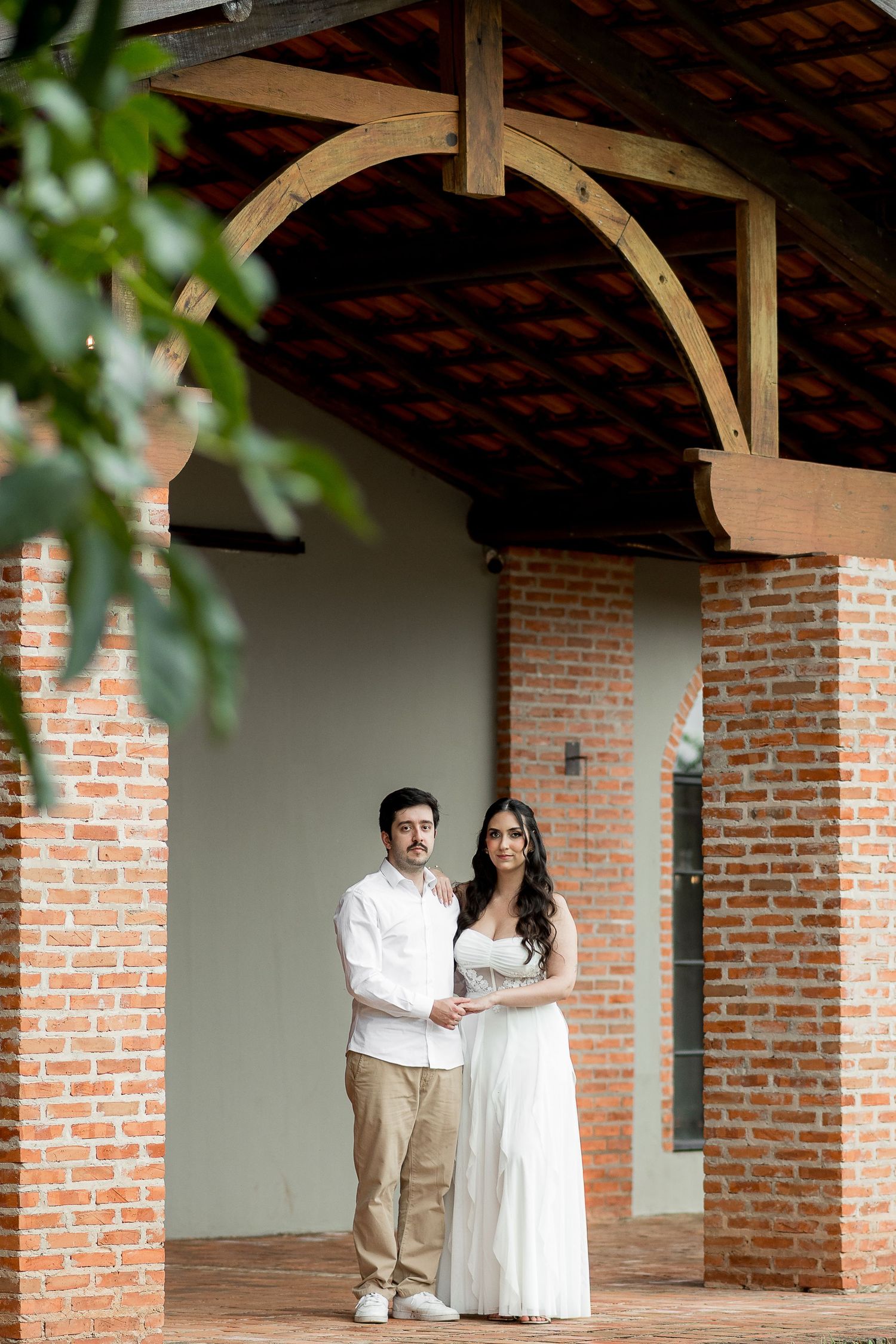 Couple in white attire posing under a rustic brick archway with wooden beams at an elegant outdoor venue.