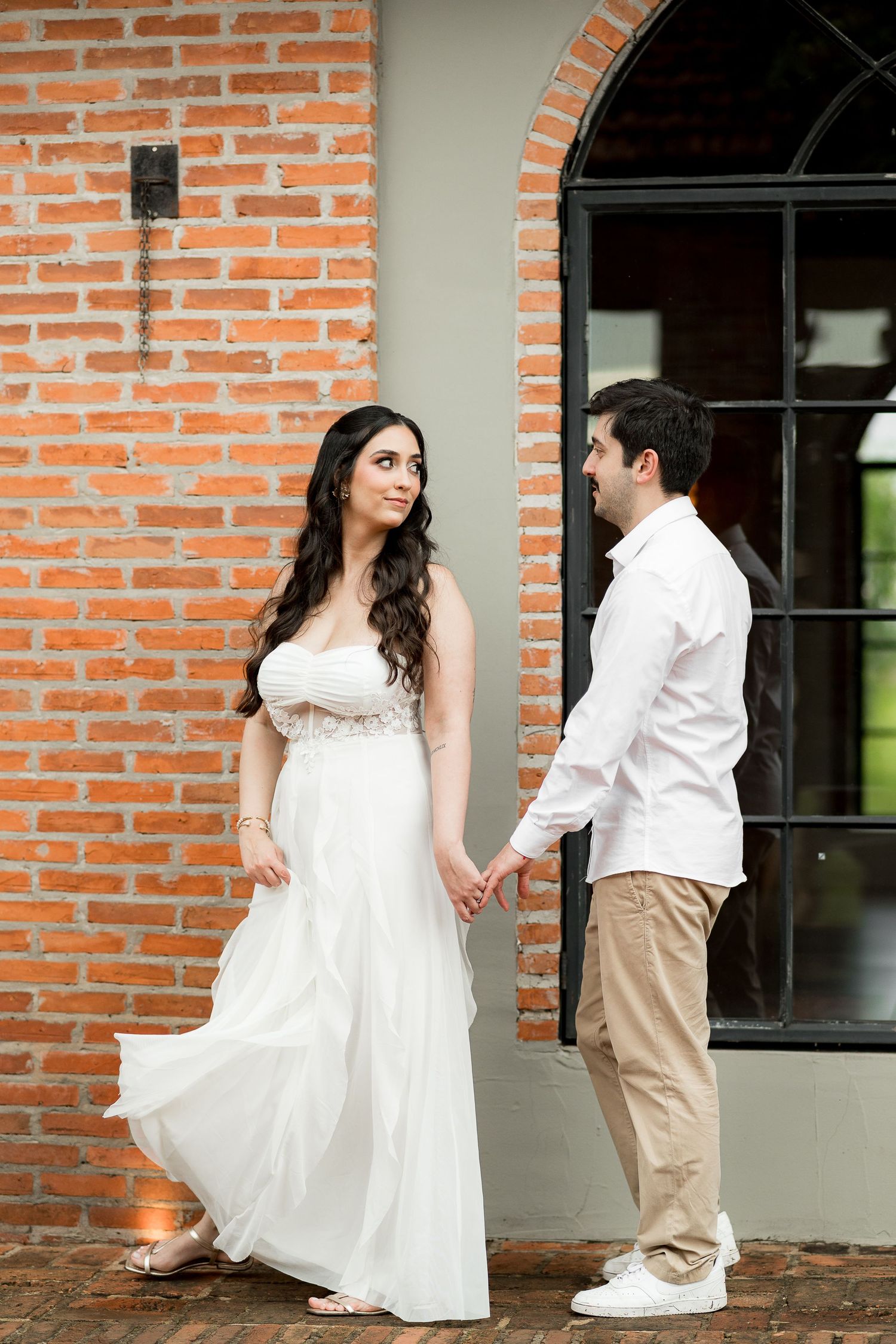 Couple in white attire holding hands by a brick wall, woman in flowing gown, man in white shirt and khakis.