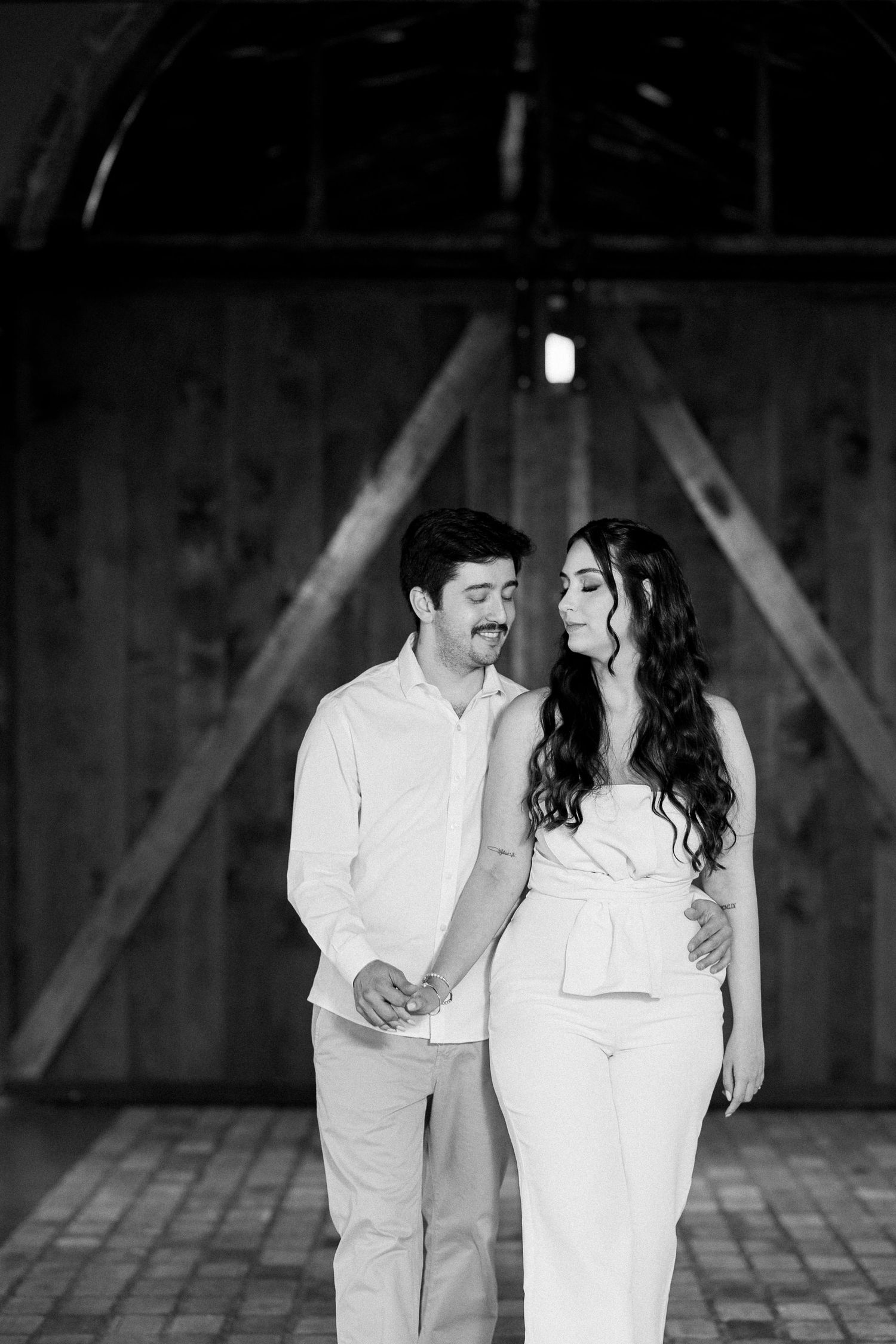 Black and white photo of a couple holding hands and gazing at each other inside a rustic barn.