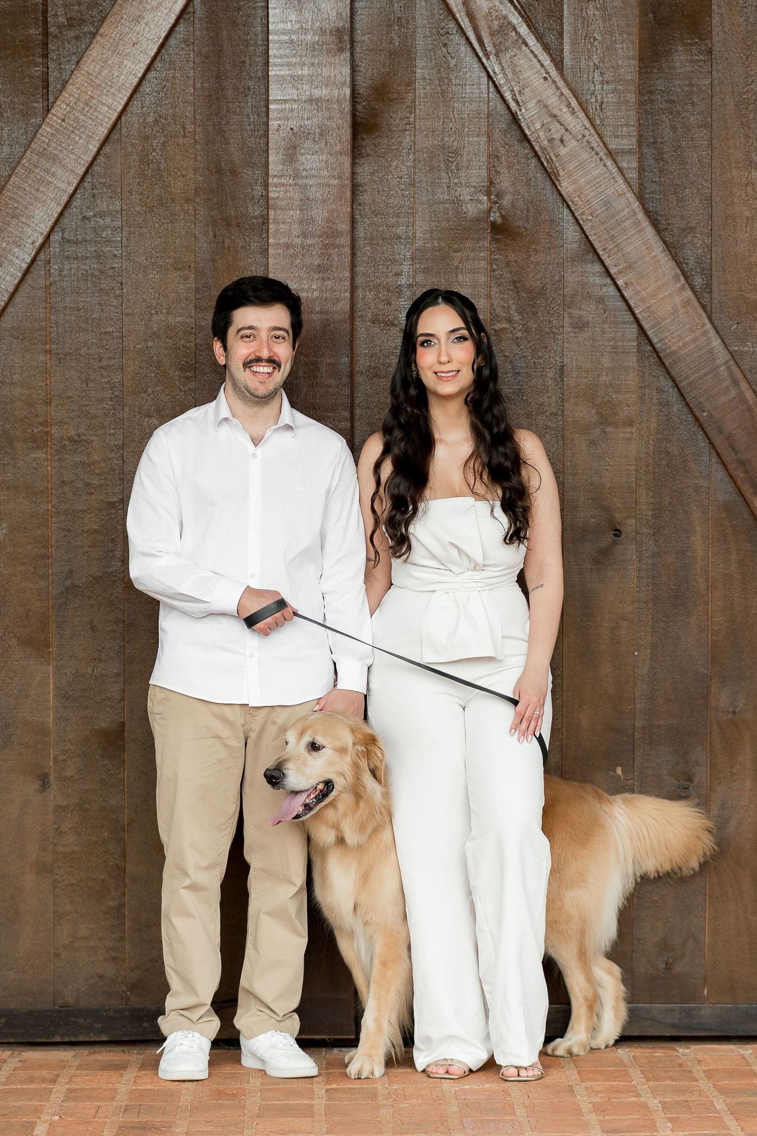Smiling couple in white outfits posing with a golden retriever on a leash in front of rustic wooden barn doors.