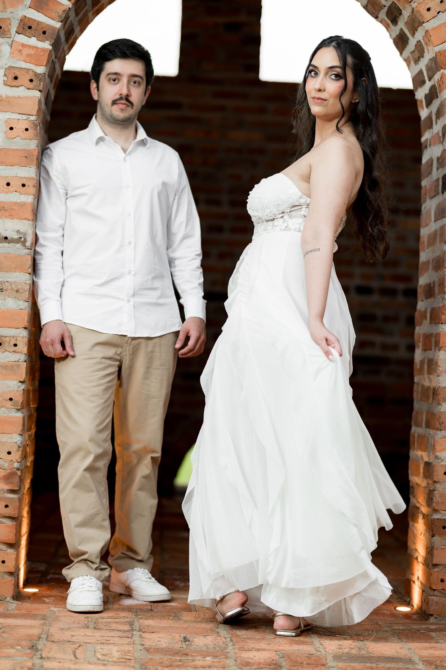A couple in white wedding attire poses in a brick archway, the bride in a strapless gown and groom in a white shirt.