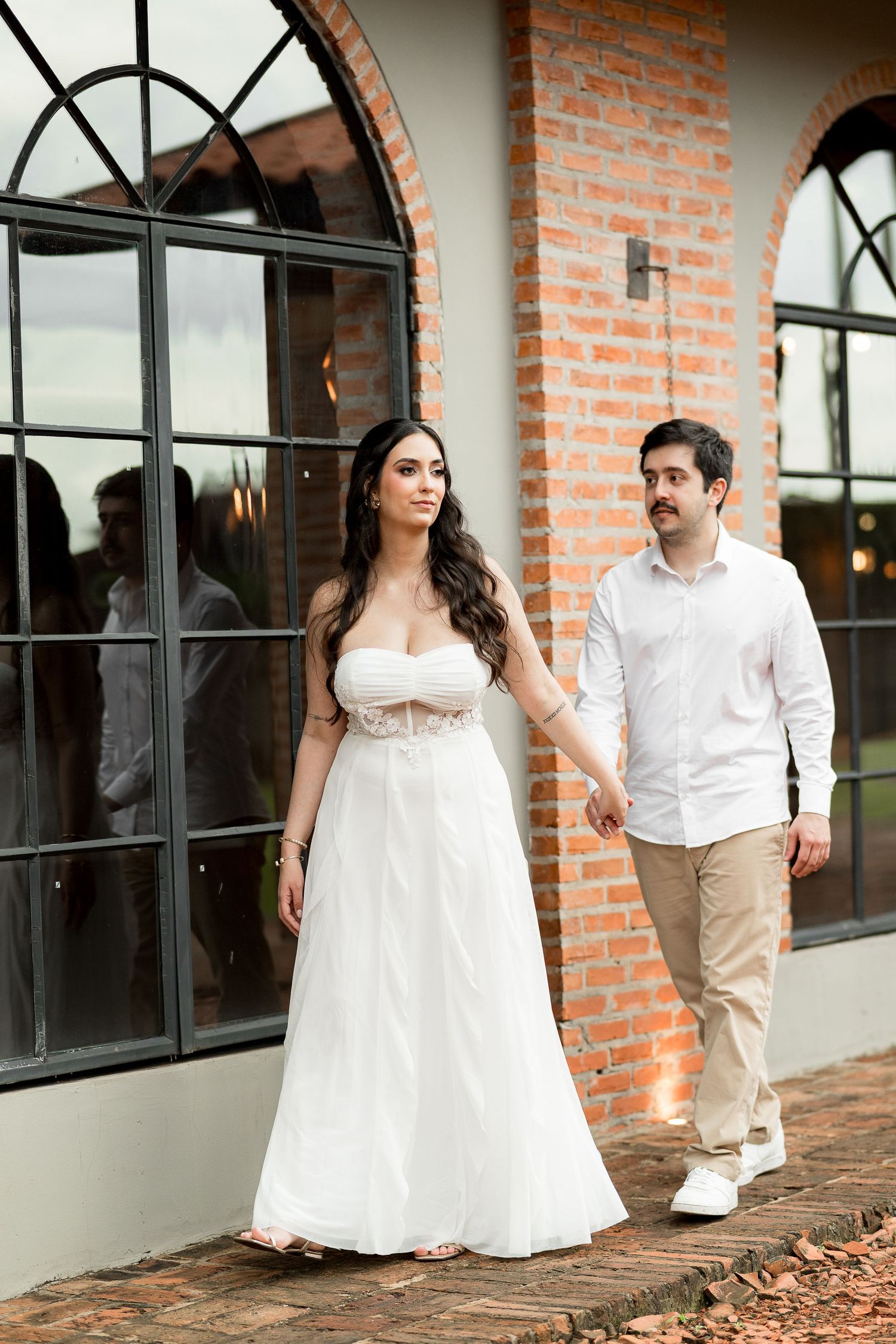 Couple in wedding attire walking hand in hand outside a brick building with arched windows.