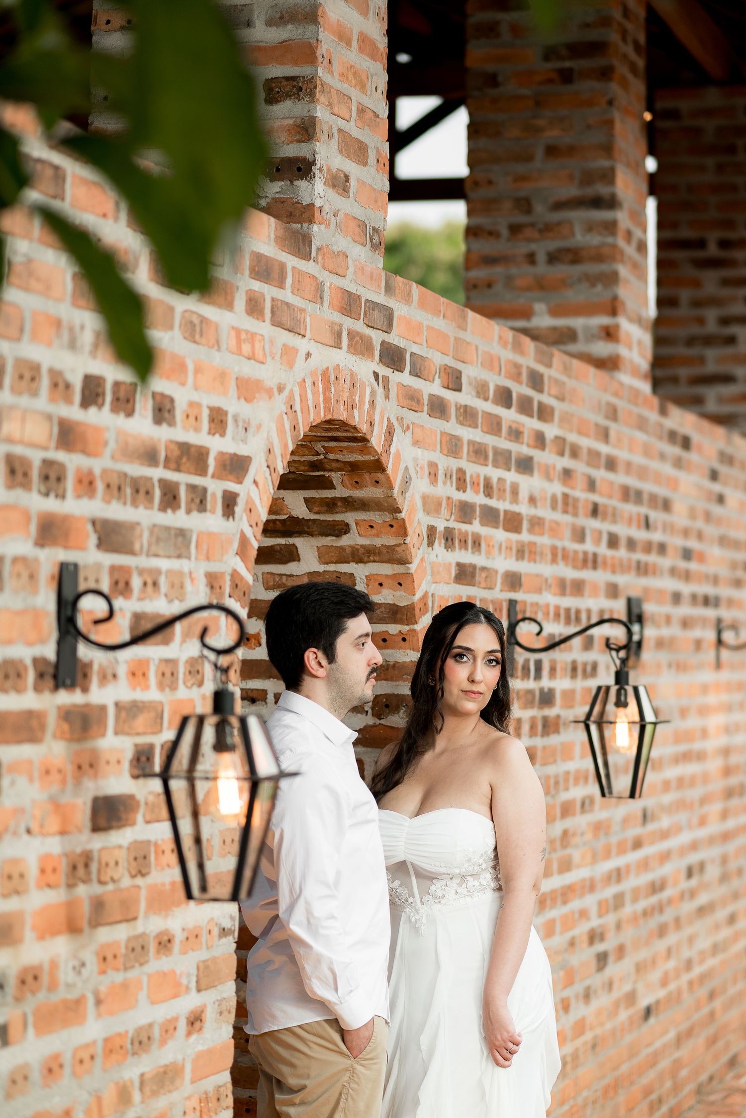 Bride and groom pose romantically against rustic brick wall with vintage lanterns in elegant outdoor wedding portrait.