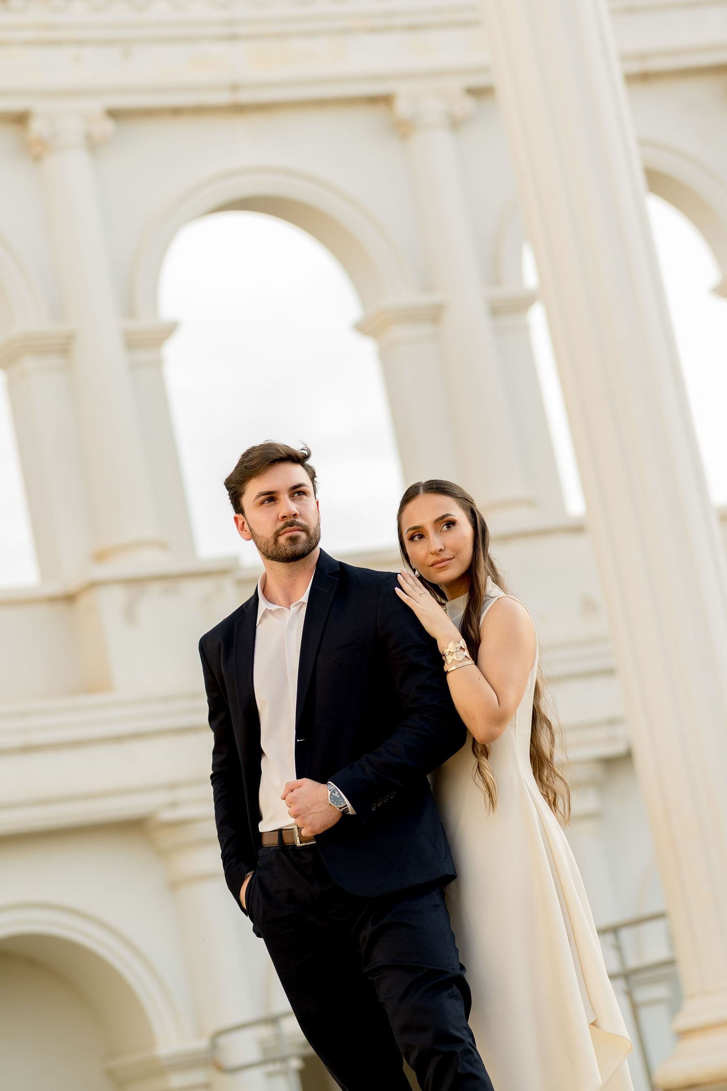 Elegant couple posing before white arched building, man in dark suit, woman in cream dress leaning on his shoulder.