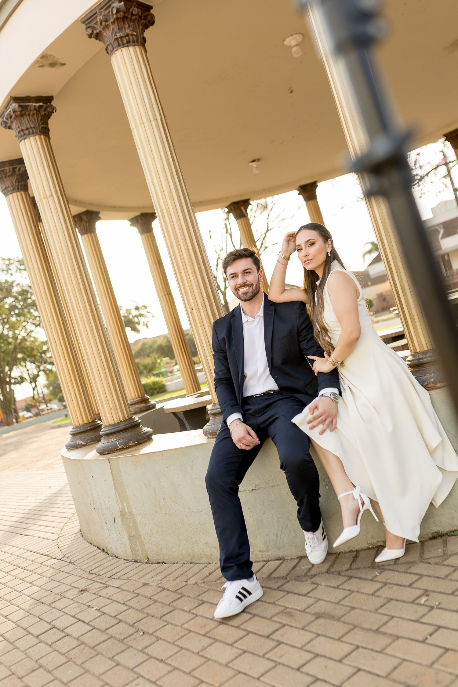 Bride and groom posing elegantly among classical columns in a park, wearing formal wedding attire.