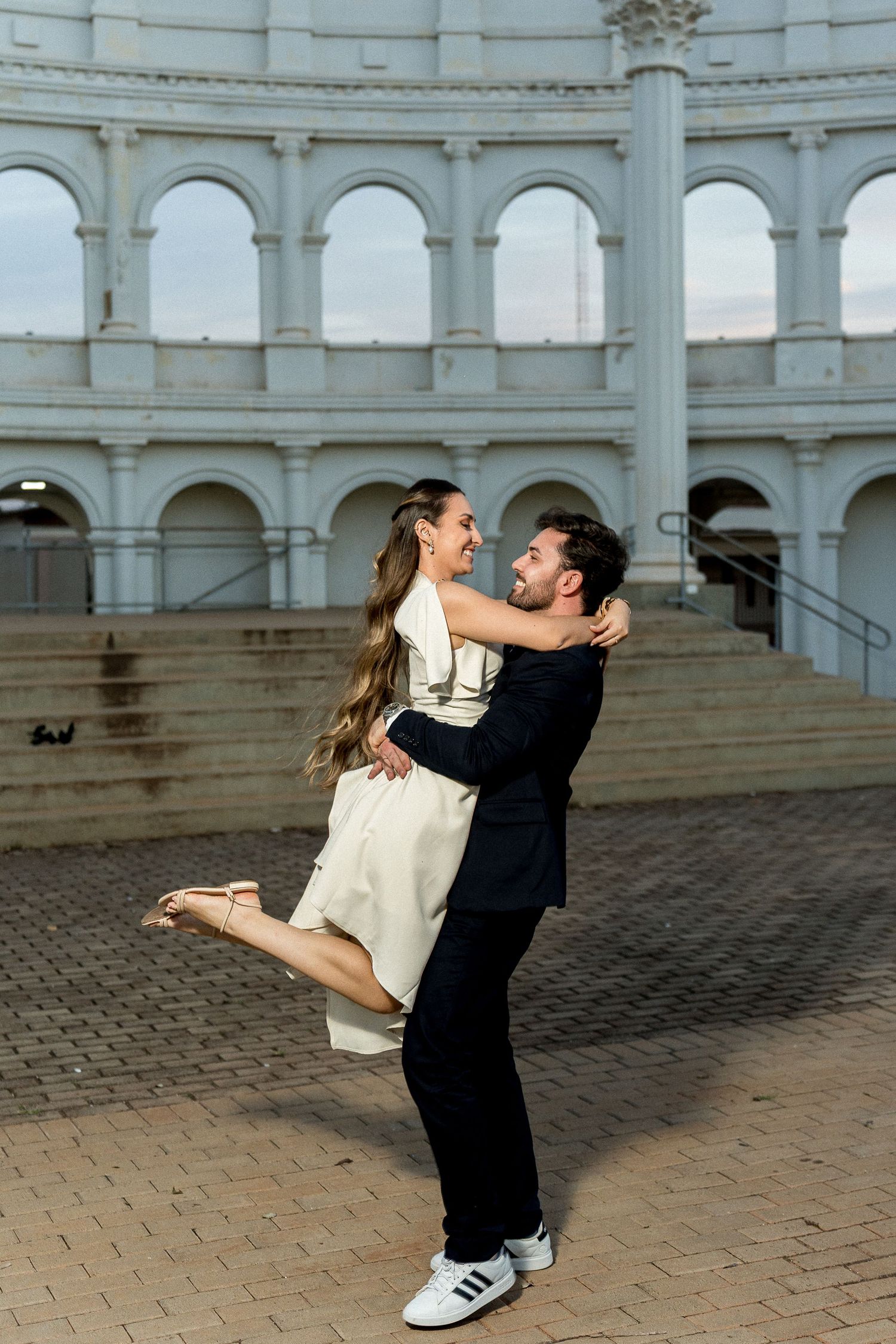 Man lifting woman in white dress, dancing joyfully in front of a grand white arched building on brick pavement.