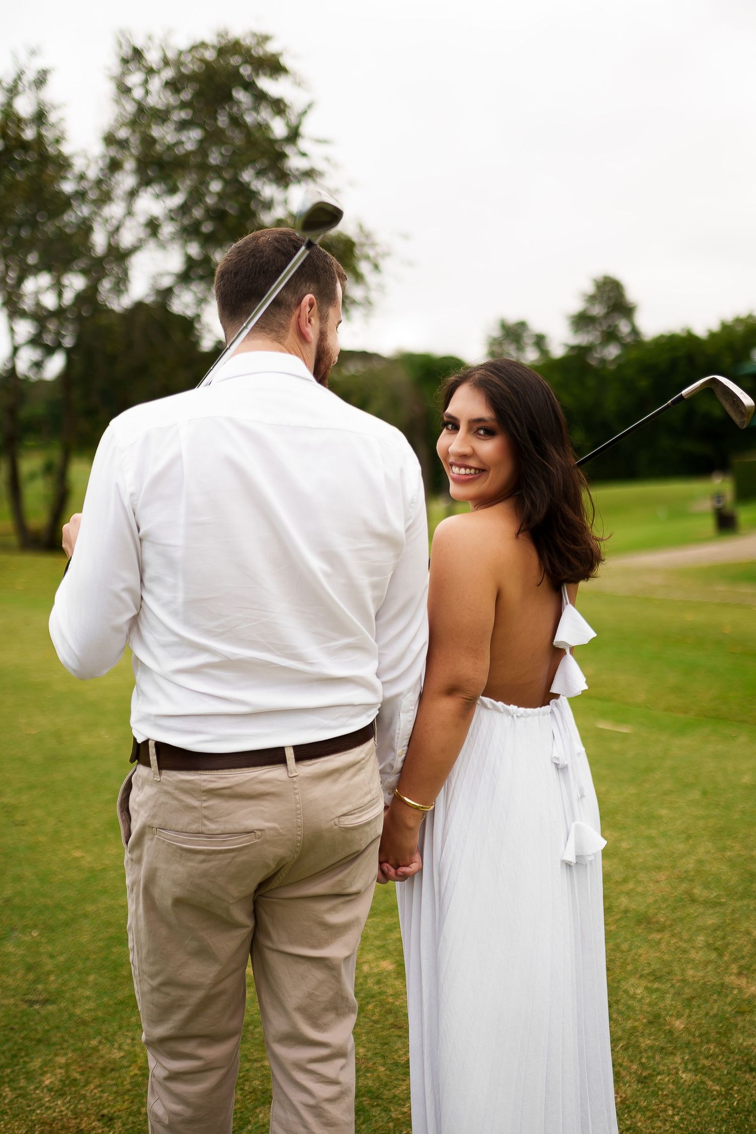 Couple holding hands on golf course, woman in white backless dress smiling, man holding golf club.