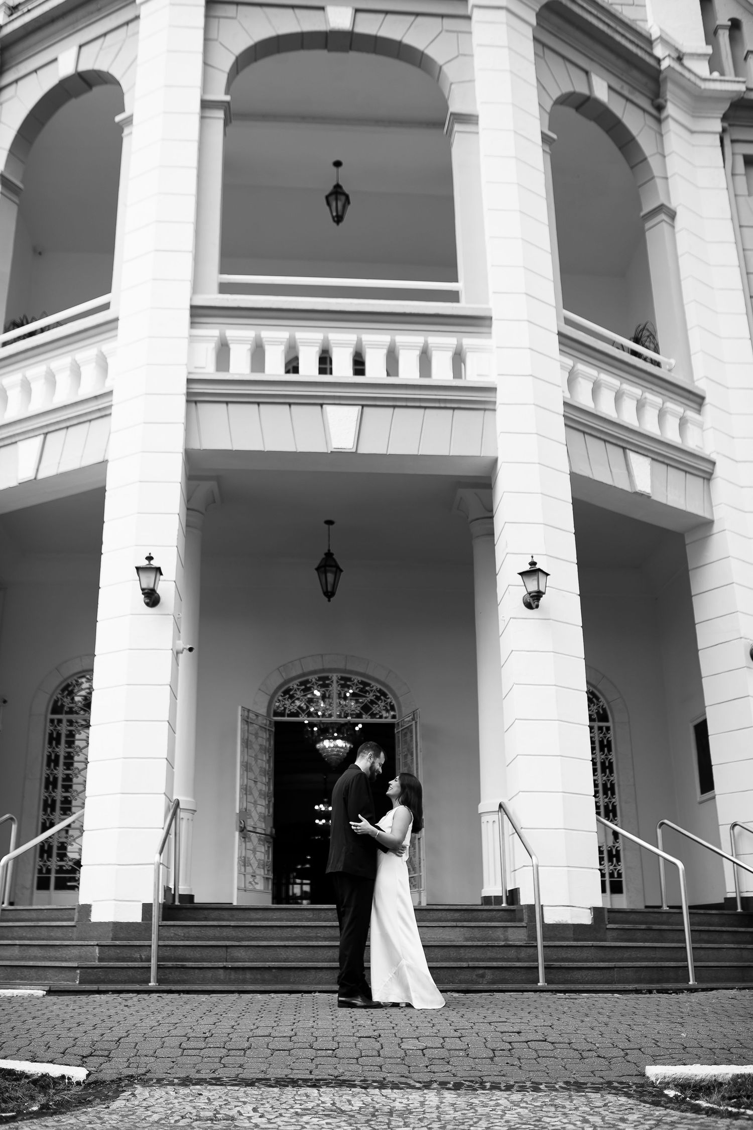 Black and white photo of a couple embracing on steps of an elegant white colonial building with arched columns.