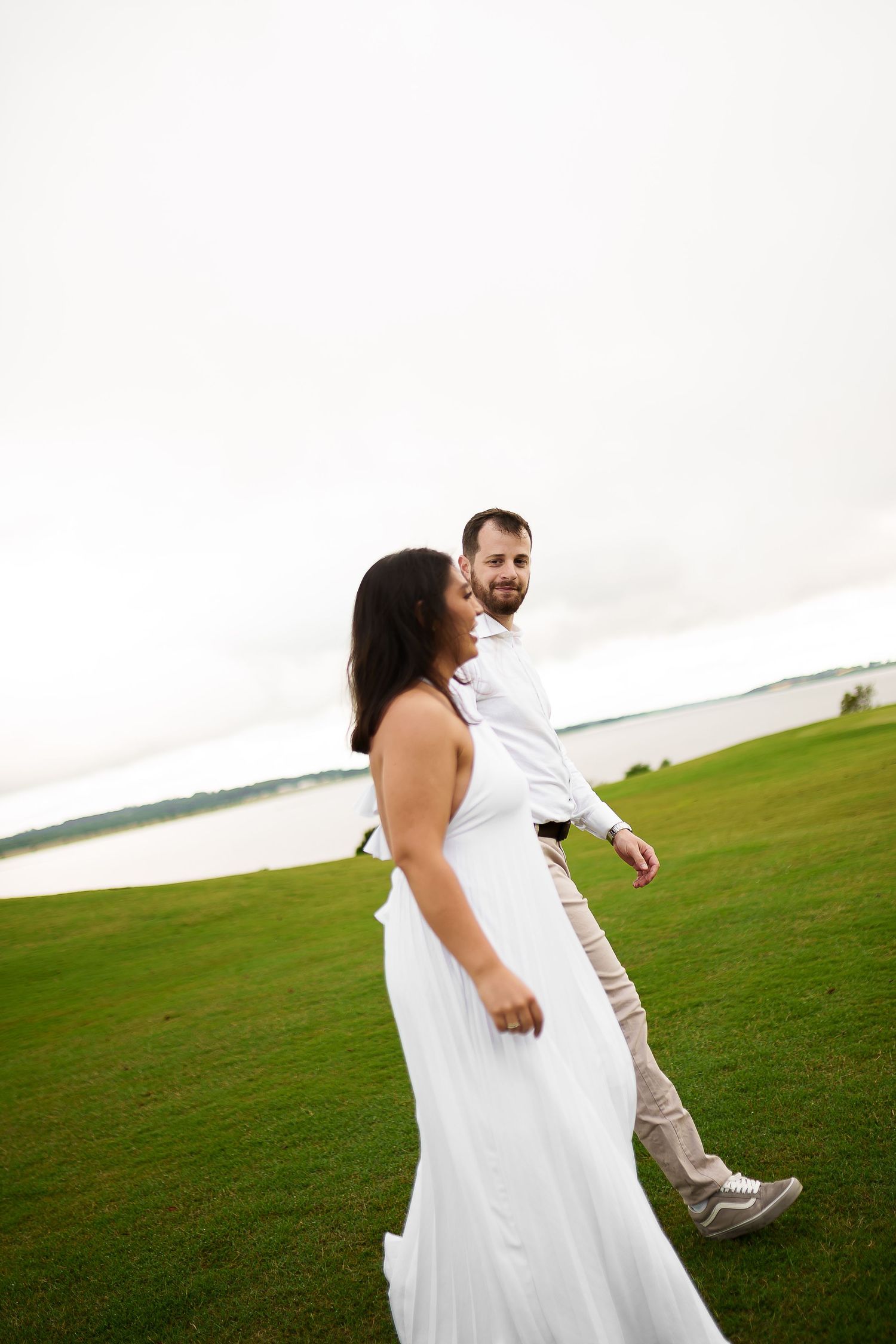 A bride in a white gown and groom in white shirt walk hand in hand on green grass near a lake.