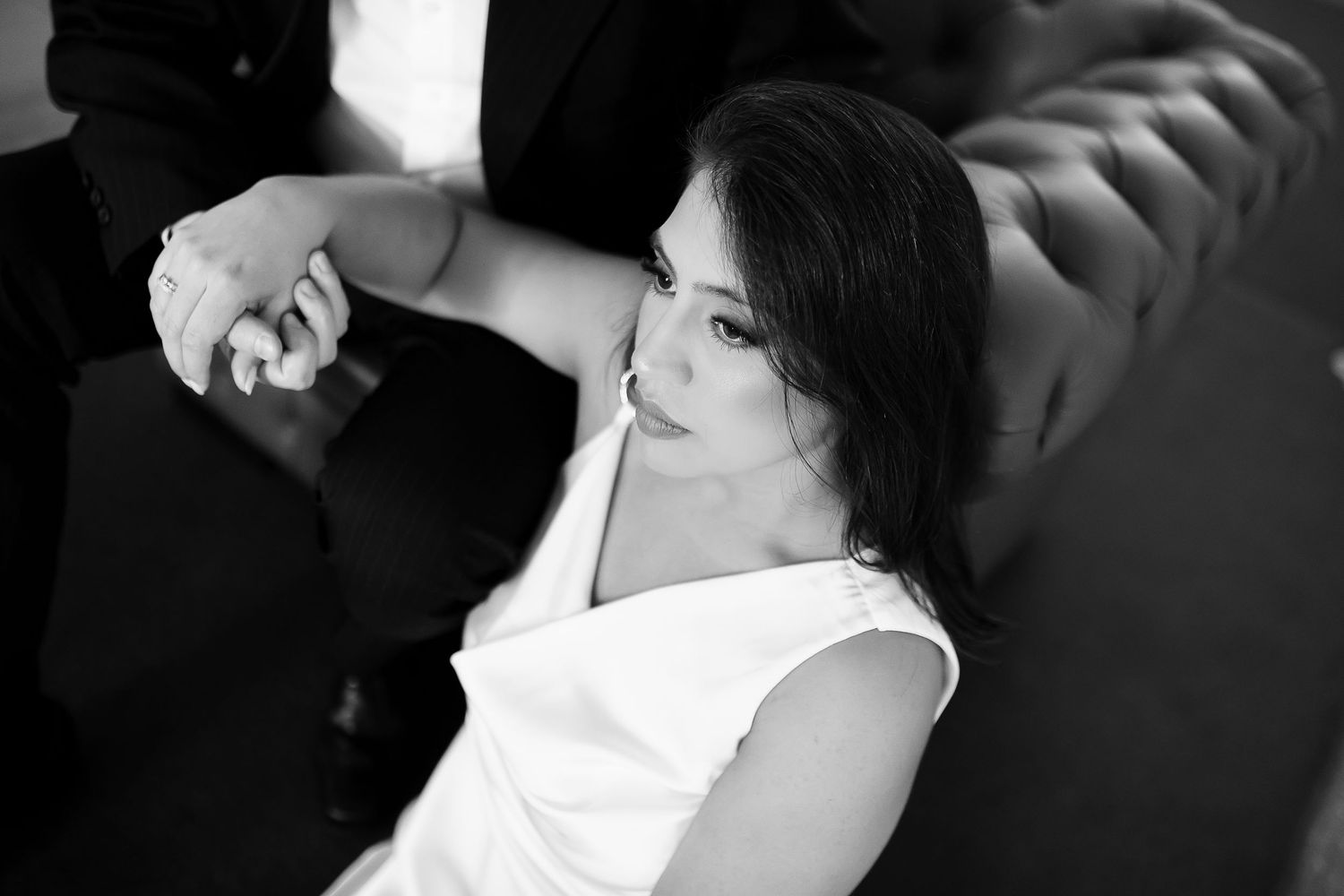 Black and white photo of an elegant woman in white dress reclining on a tufted sofa, holding a mans hand.