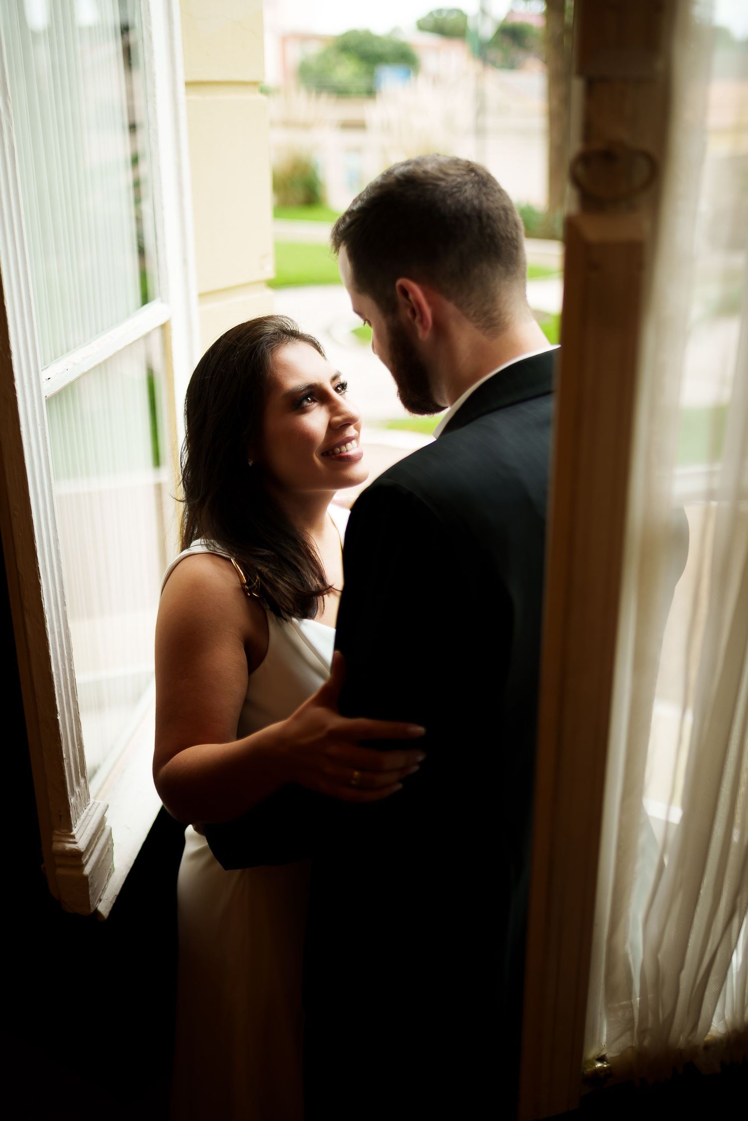 A smiling bride in white dress gazes at groom in black suit near an open window in romantic setting.