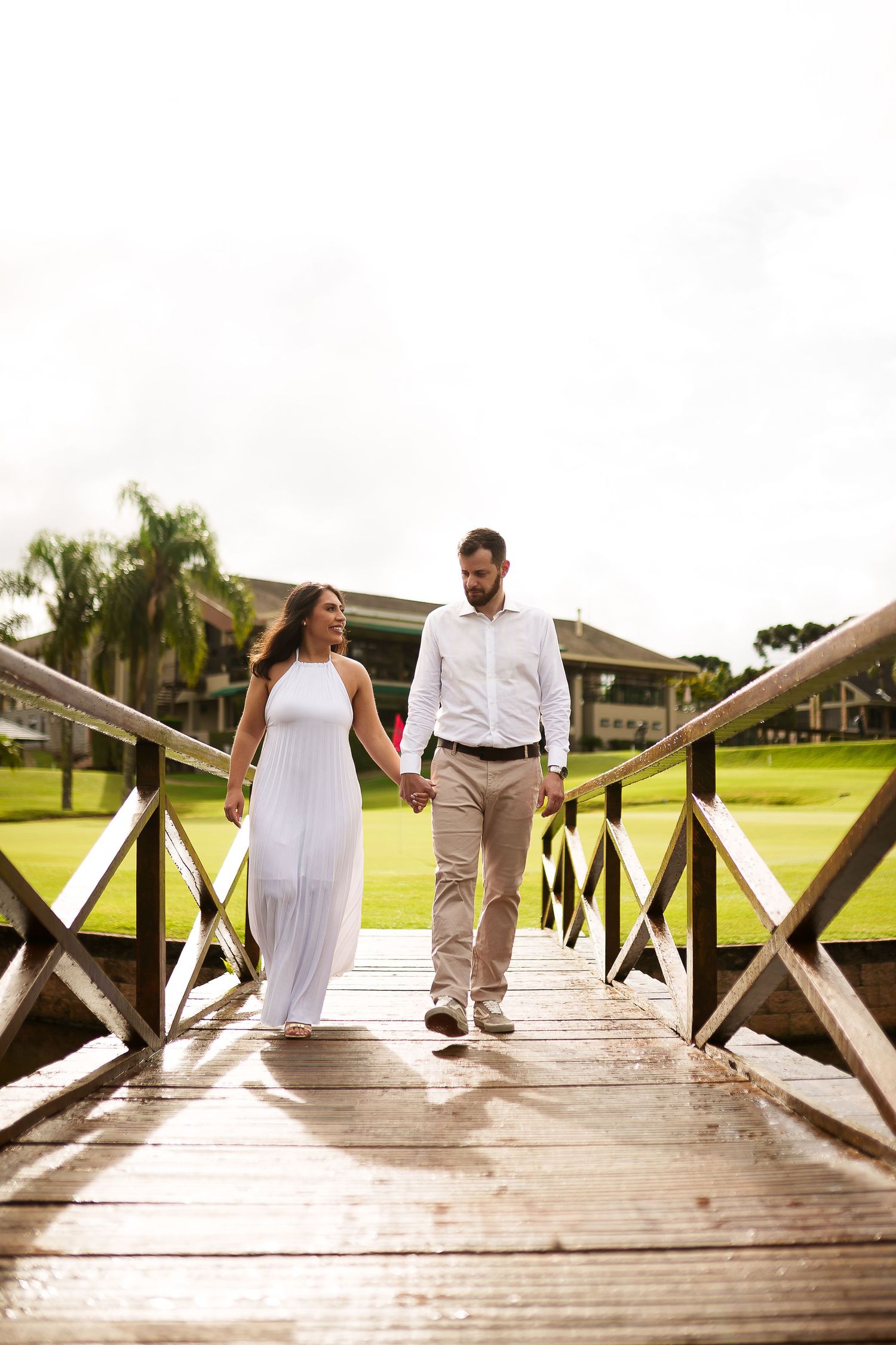 Couple in white dress and casual attire hold hands walking on wooden bridge outdoors.