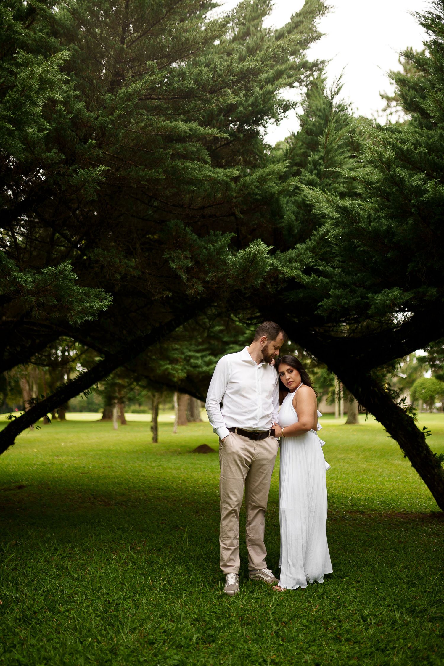 Couple embracing in lush green park under arching trees, man in white shirt and woman in white dress.