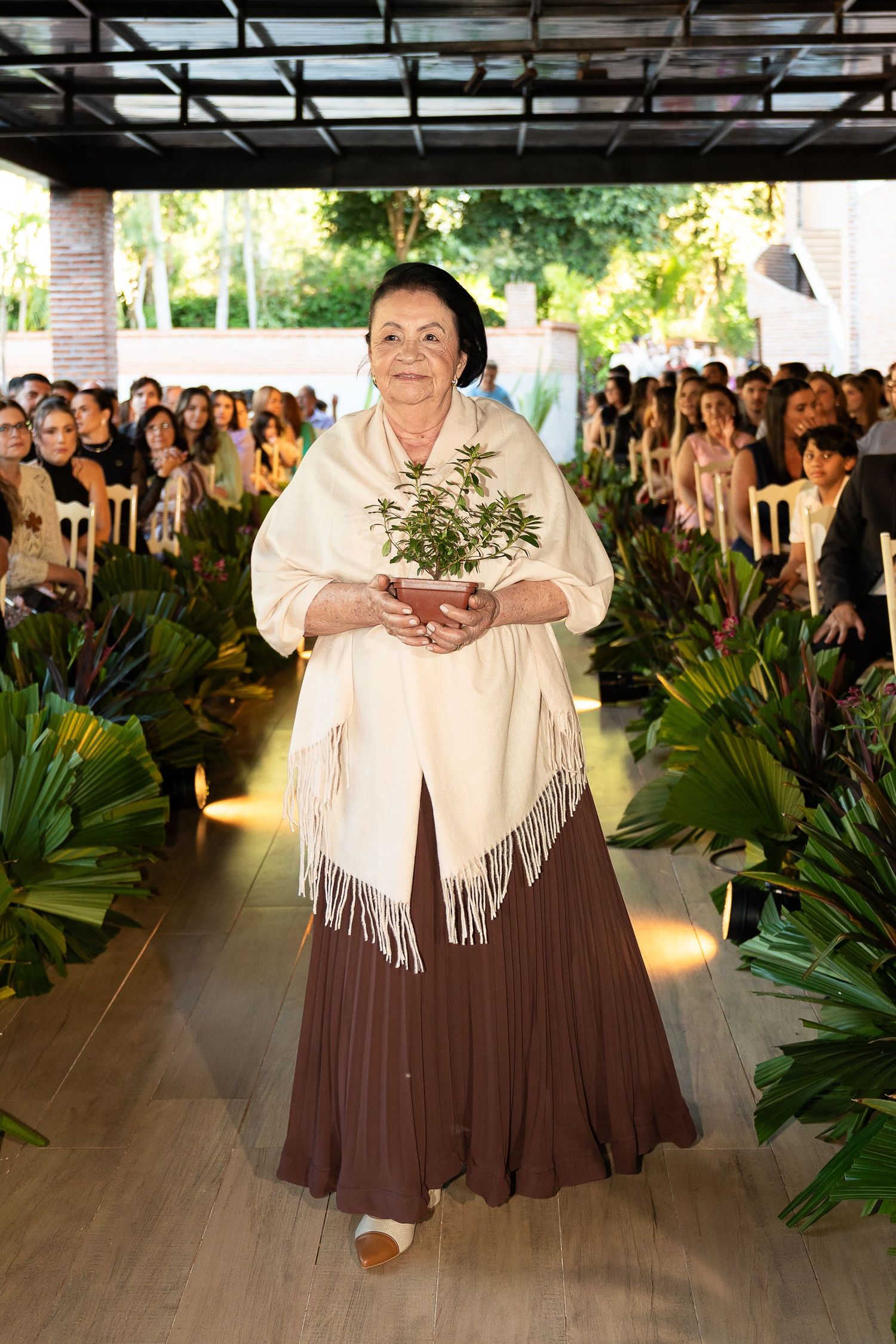 Elderly woman in cream shawl and brown skirt walks down aisle carrying potted plant at outdoor wedding ceremony.