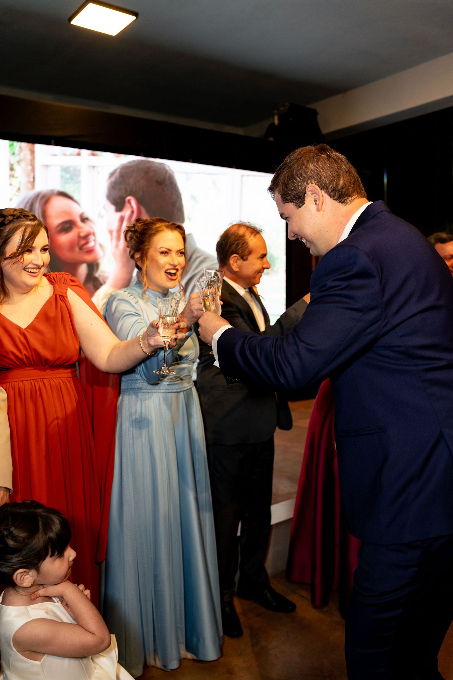 Guests in formal attire toast with champagne glasses at an elegant wedding reception with a large screen backdrop.