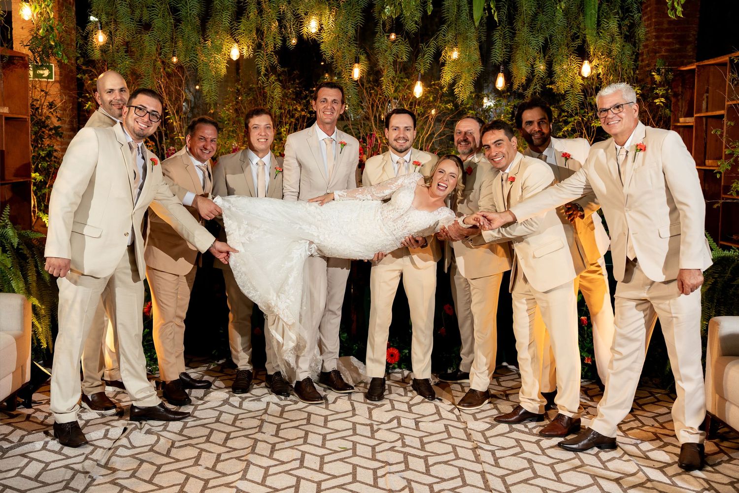 Groom and groomsmen in beige suits lifting bride in white wedding dress outdoors at elegant evening reception.