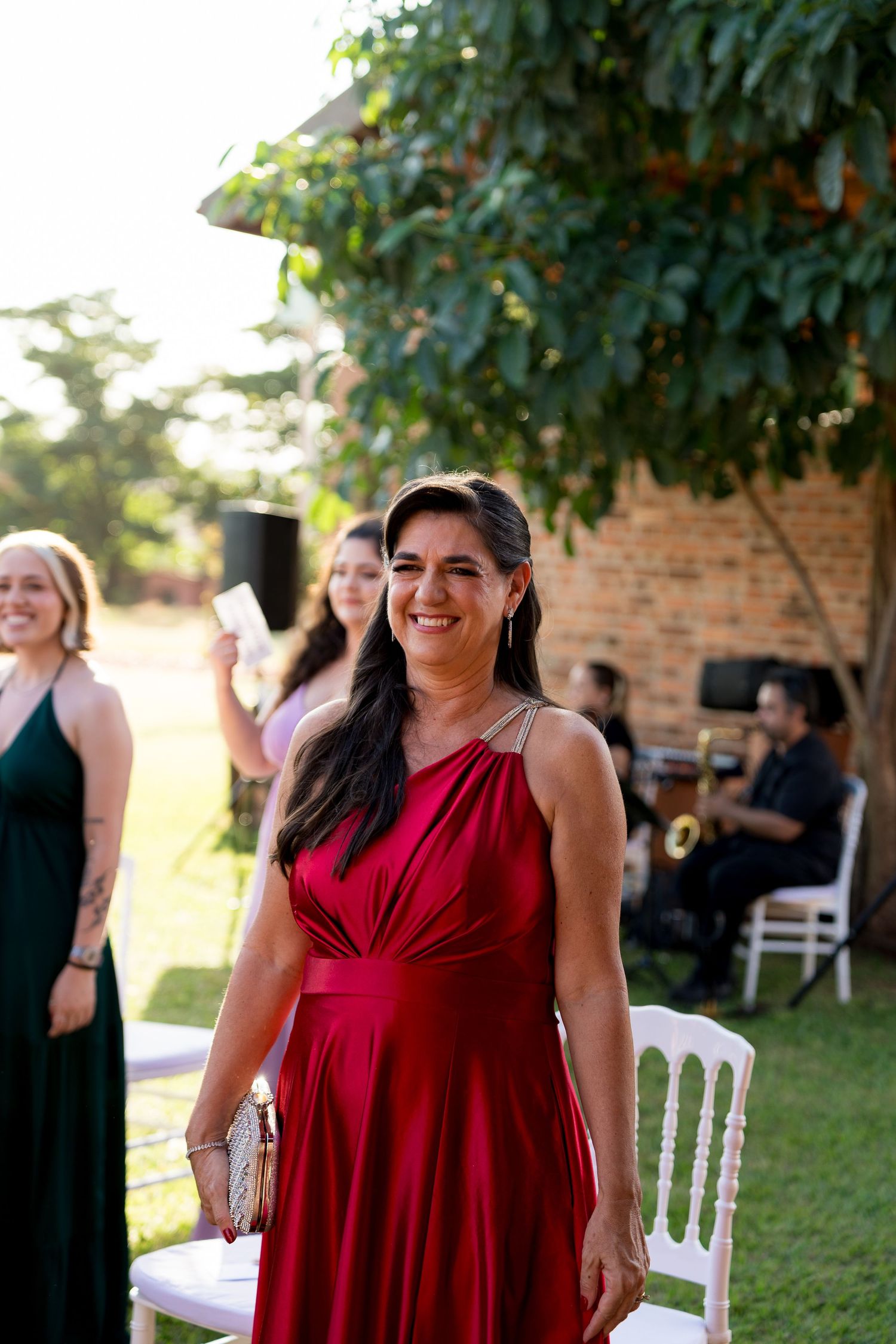 Woman in red gown smiling at outdoor wedding ceremony with guests and live band in background.