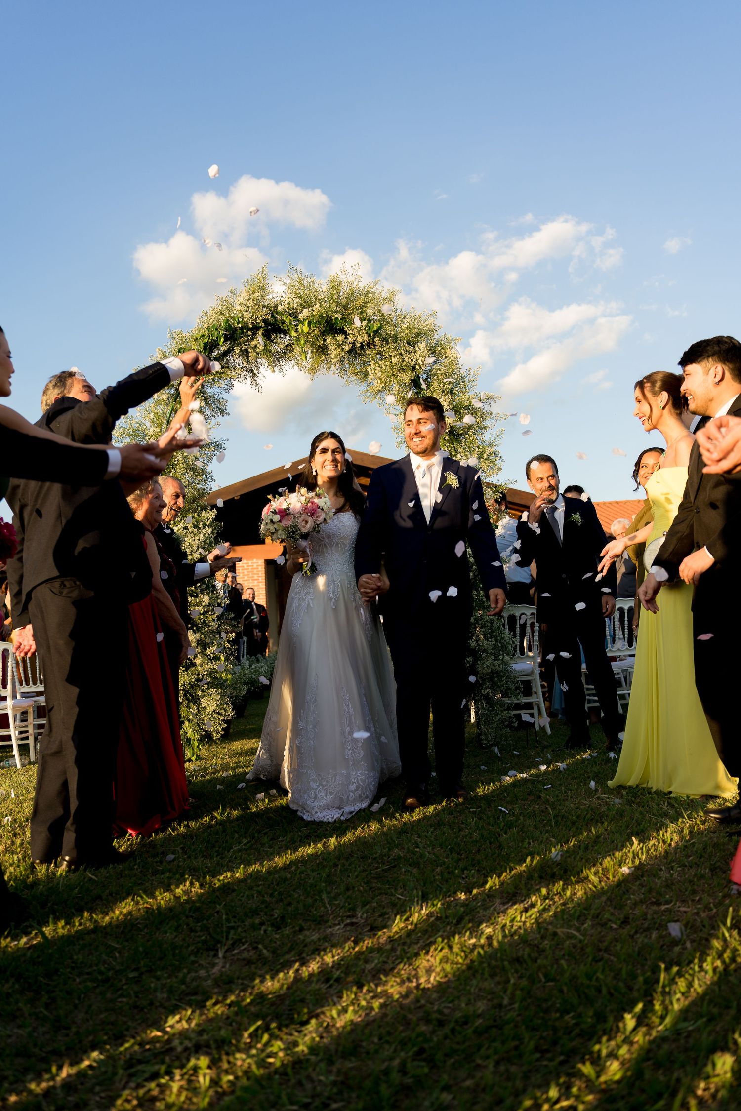 Newlywed couple walks down outdoor aisle under floral arch while guests toss petals on sunny wedding day.