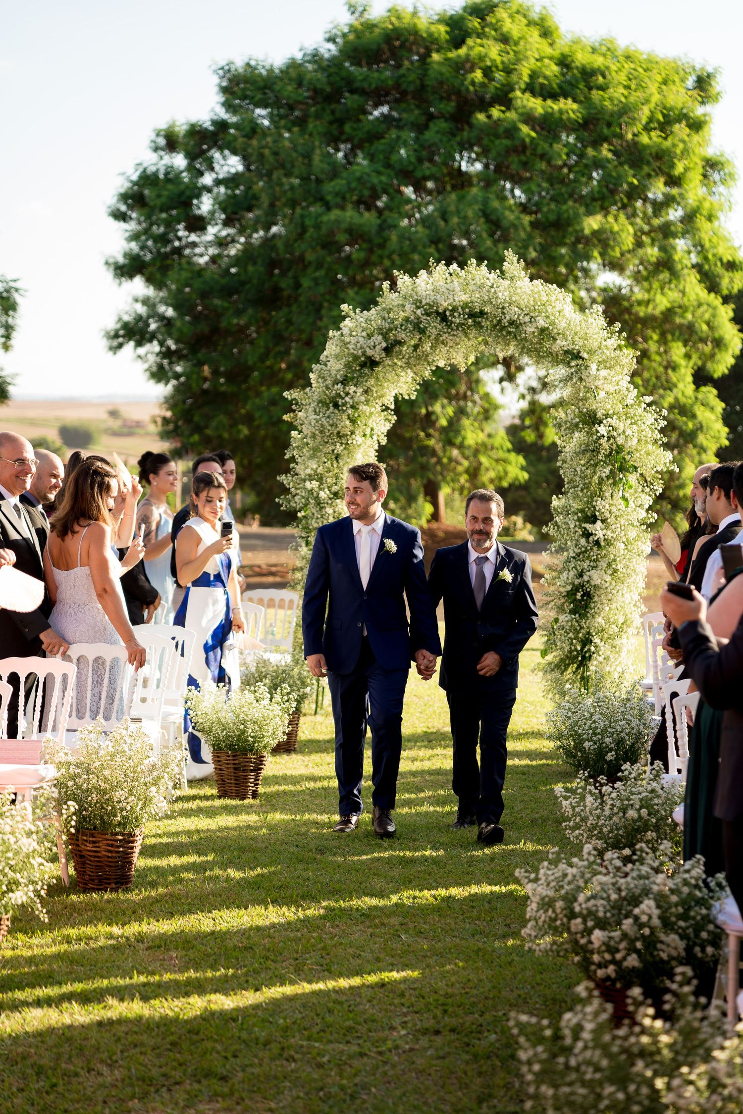 Two grooms walk hand-in-hand down a floral arch aisle at an outdoor wedding ceremony surrounded by guests.