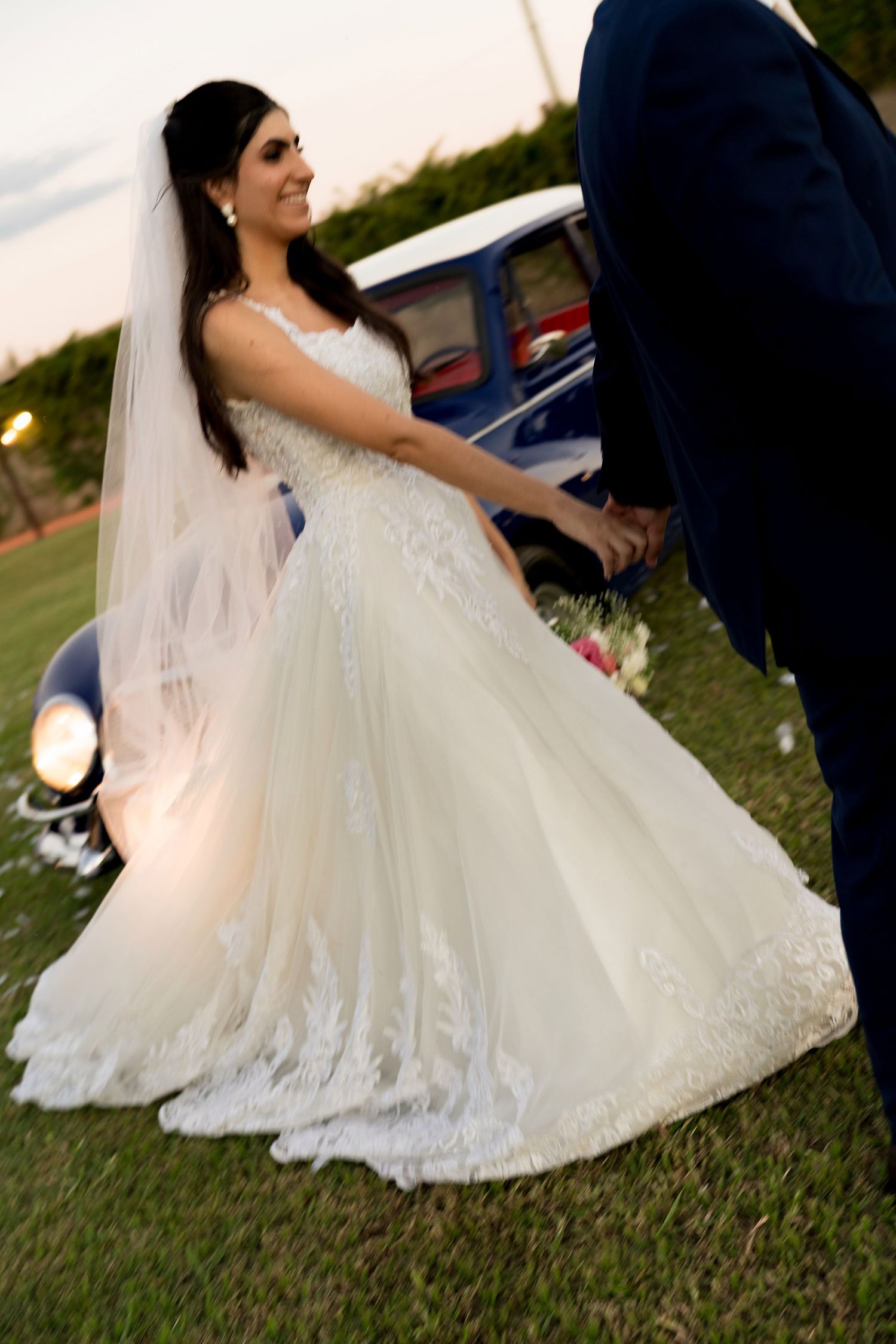 Bride in lace ball gown wedding dress with veil holds groom's hand outdoors near vintage blue car at sunset.