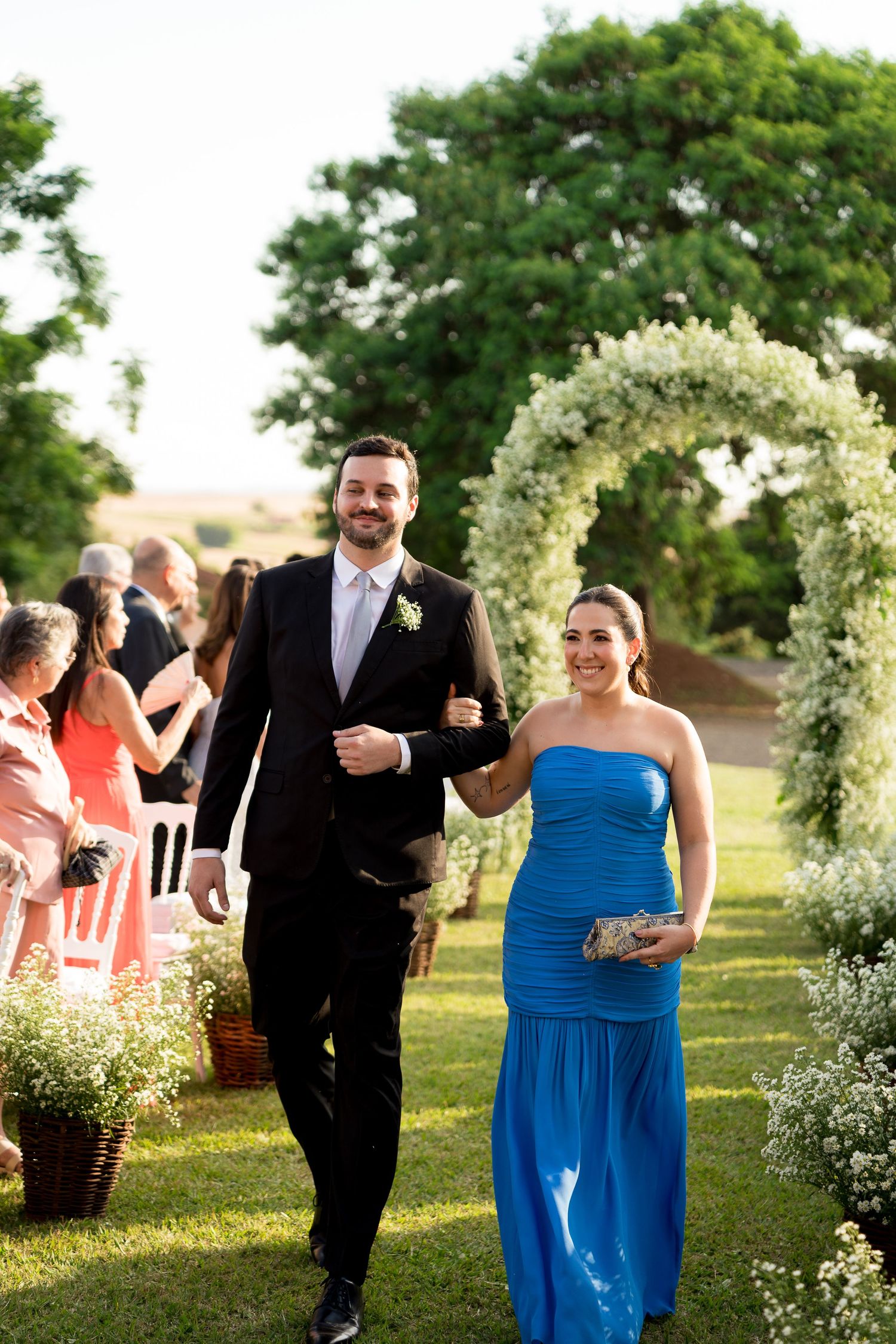 A smiling couple walks arm in arm down an outdoor wedding aisle, man in black suit, woman in blue strapless gown.