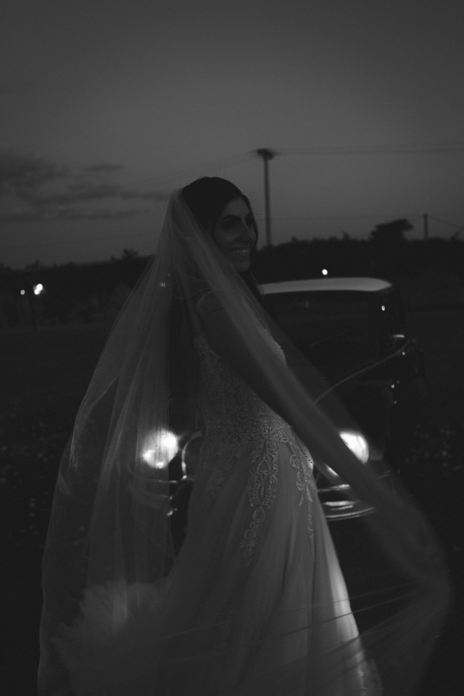 Black and white photo of a bride in a lace gown and veil standing by a vintage car at dusk.