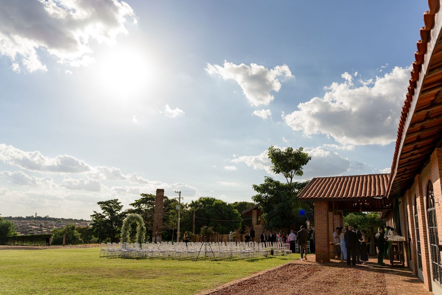 Outdoor wedding venue with white chairs on green lawn beside rustic building under bright sunny sky with clouds.