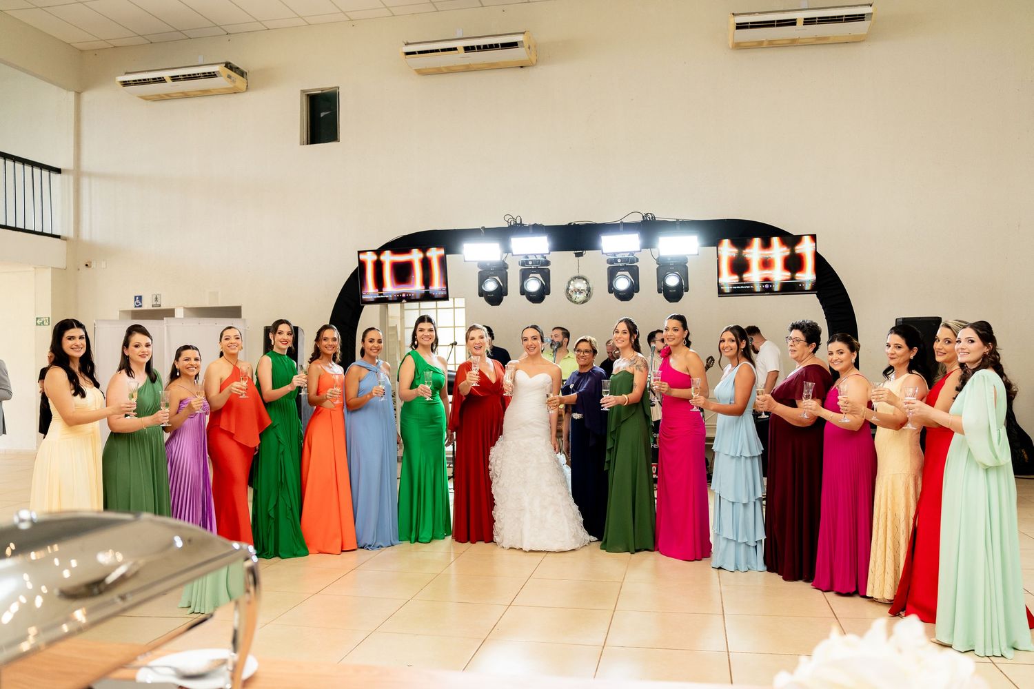 Bride in white gown poses with colorful bridesmaids in rainbow dresses at elegant wedding reception hall.