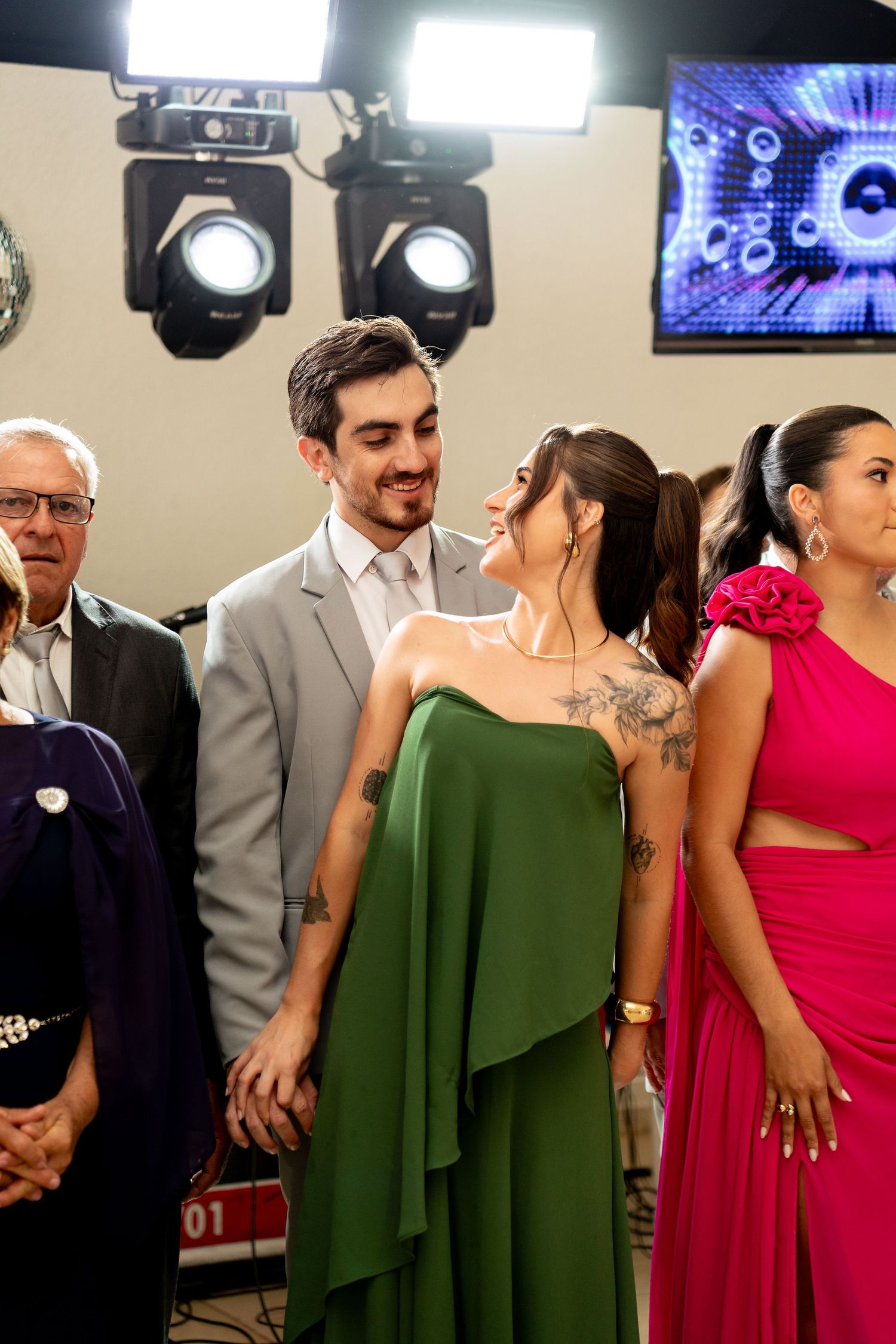 Elegant couple in green and gray formal attire smiling at an event, with guests in colorful gowns and stage lights behind.