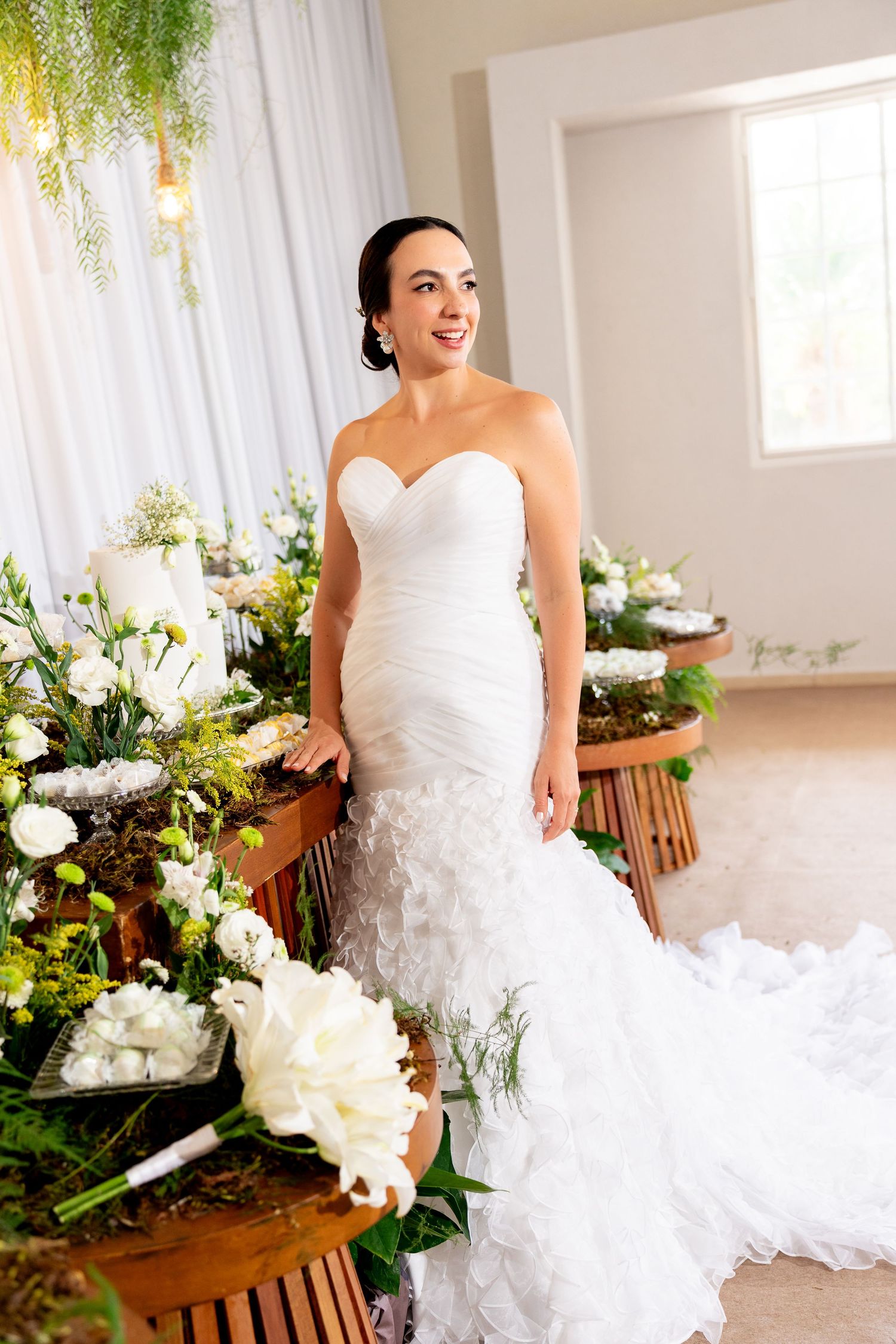 Bride in strapless mermaid wedding gown poses among lush white floral arrangements in an elegant venue.