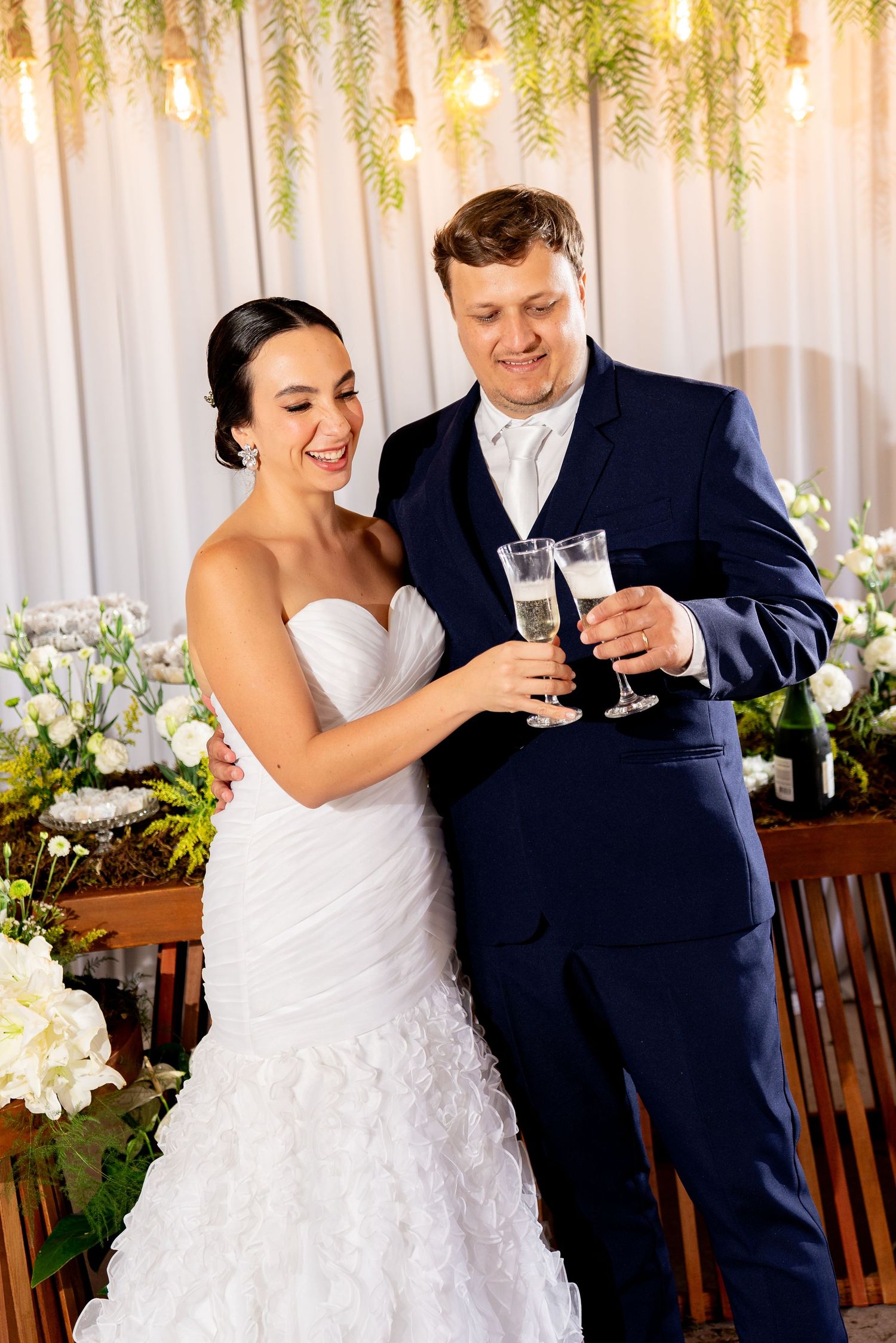 A smiling bride in a white gown and groom in navy suit toasting with champagne flutes at their wedding reception.