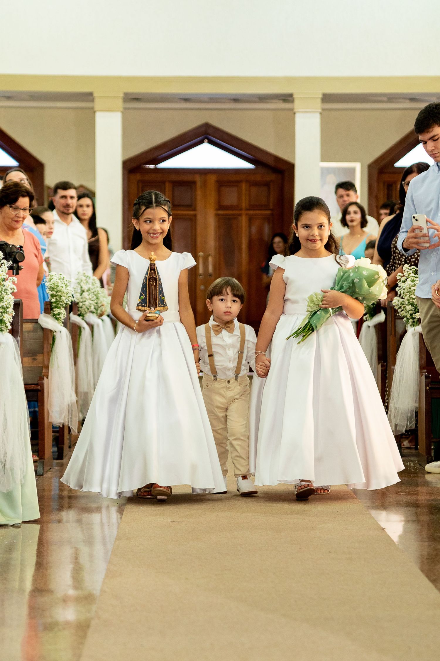 Two flower girls in white dresses and a ring bearer walk down a church aisle during a wedding ceremony.