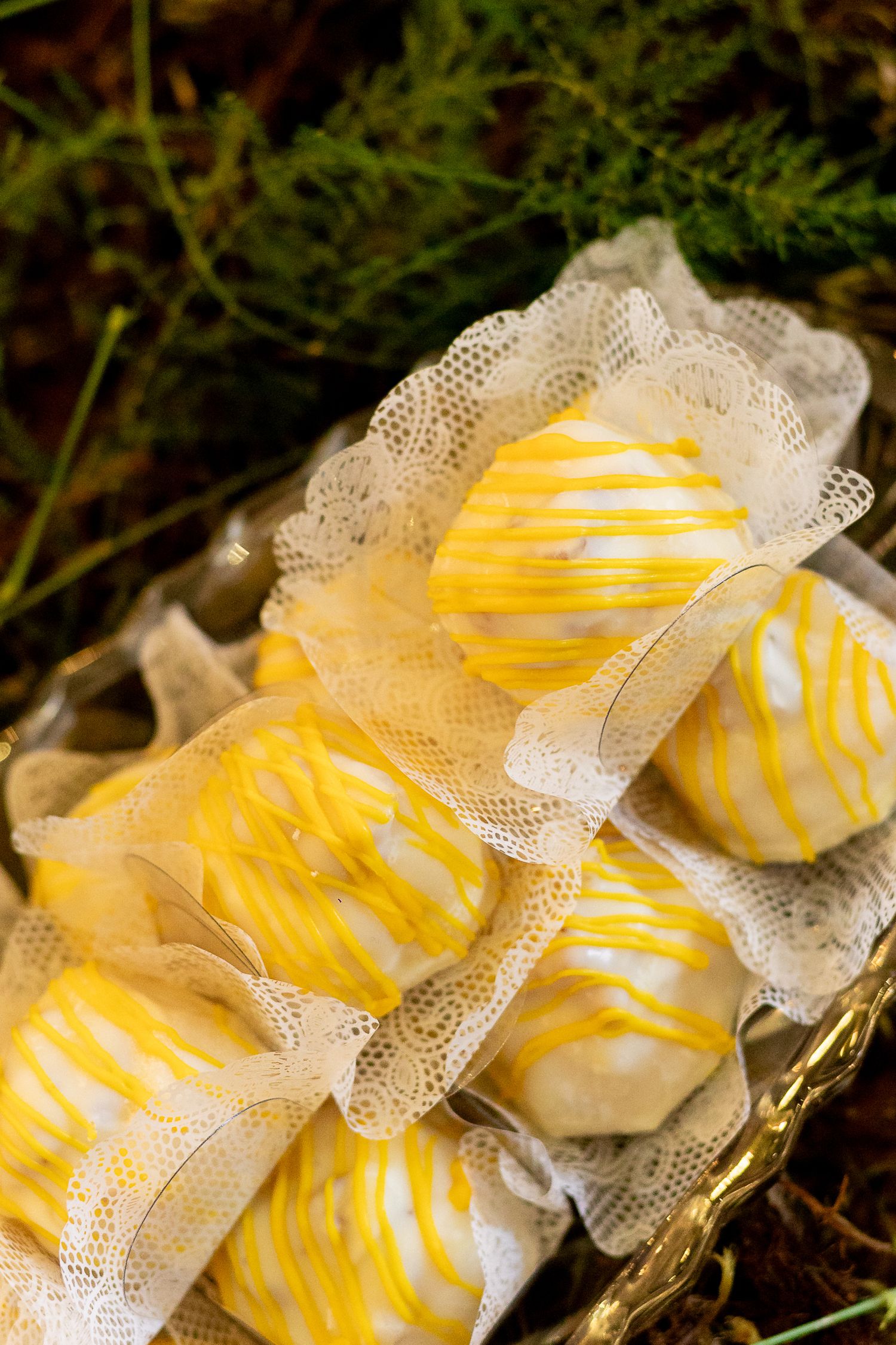 Yellow and white striped cake pops arranged in a gold basket with lace ribbon, set against green foliage.
