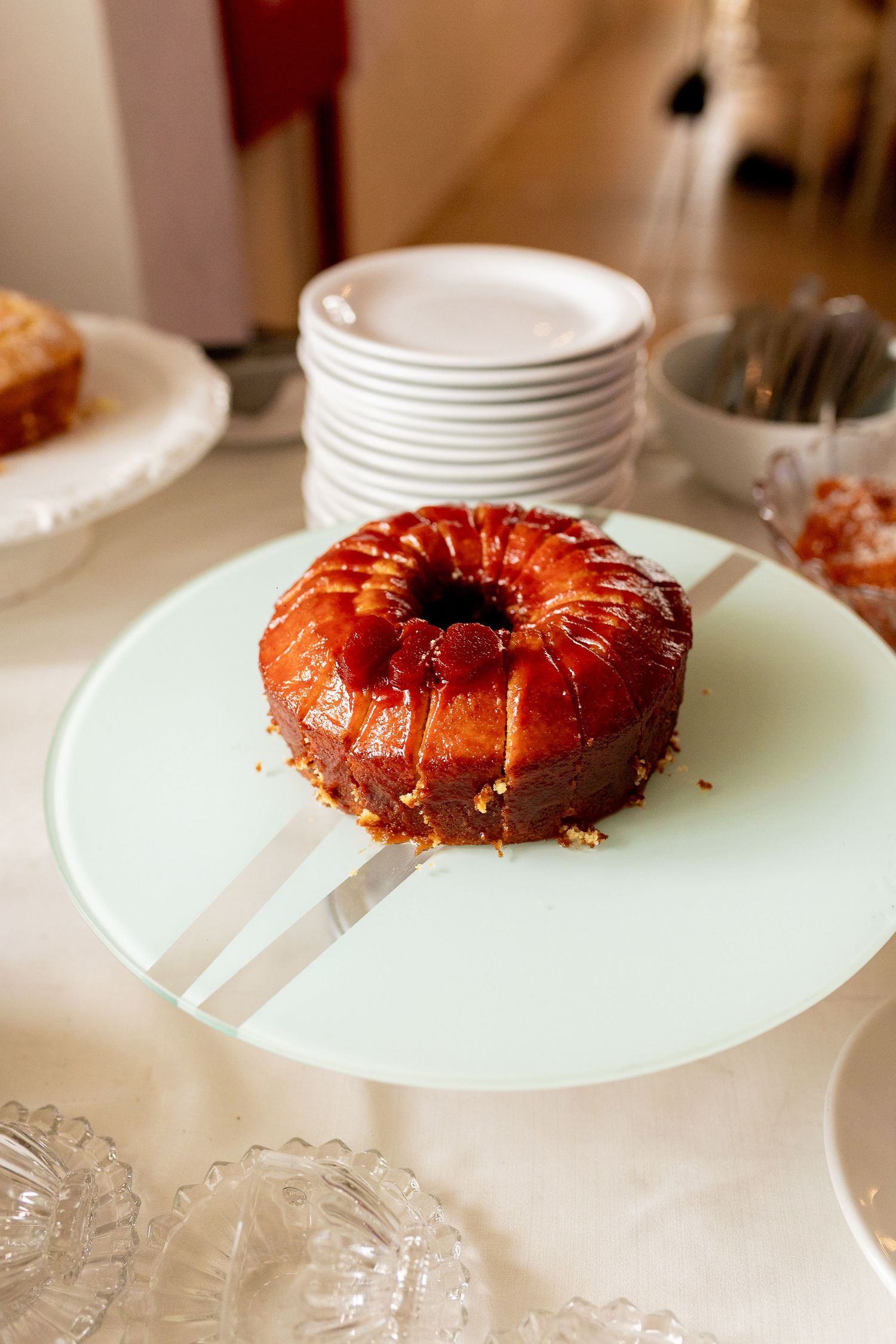 A glazed bundt cake with caramel topping displayed on a white cake stand at a dessert table.
