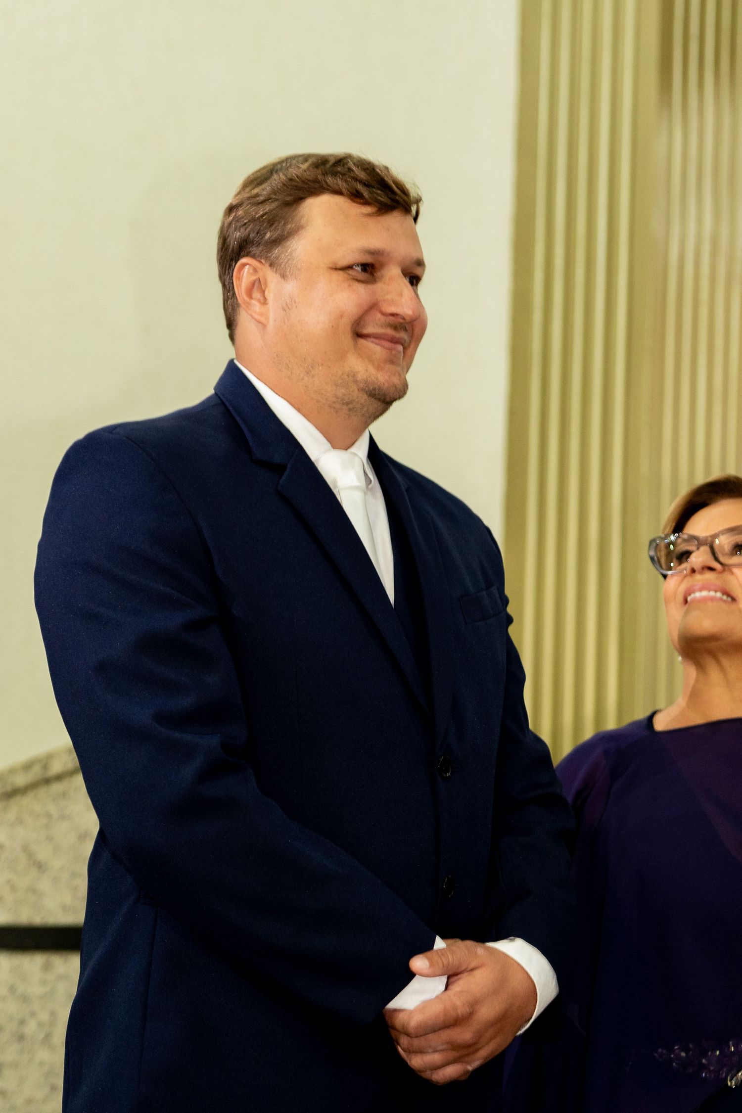 A smiling man in a navy blue suit stands beside a woman in glasses at a formal indoor event.